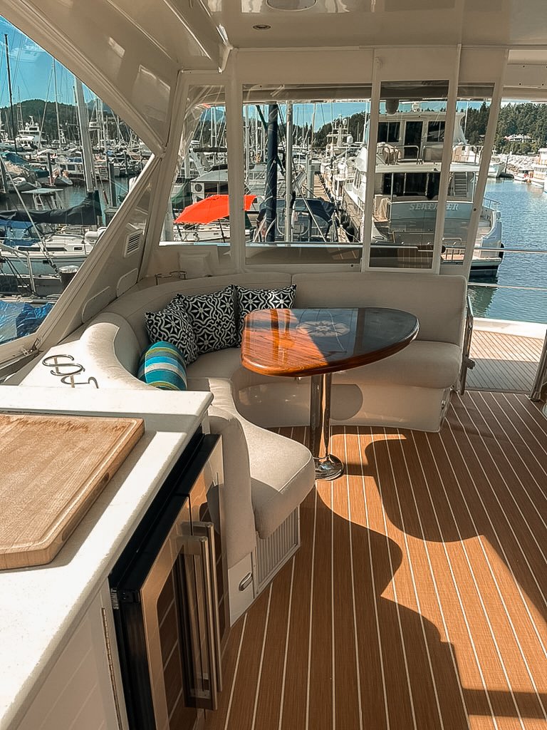 Interior of a boat with a curved white cushioned seating area, black and white patterned pillows, a striped pillow, a small round wooden table, a countertop with a cutting board, and large windows showing a marina with boats docked.