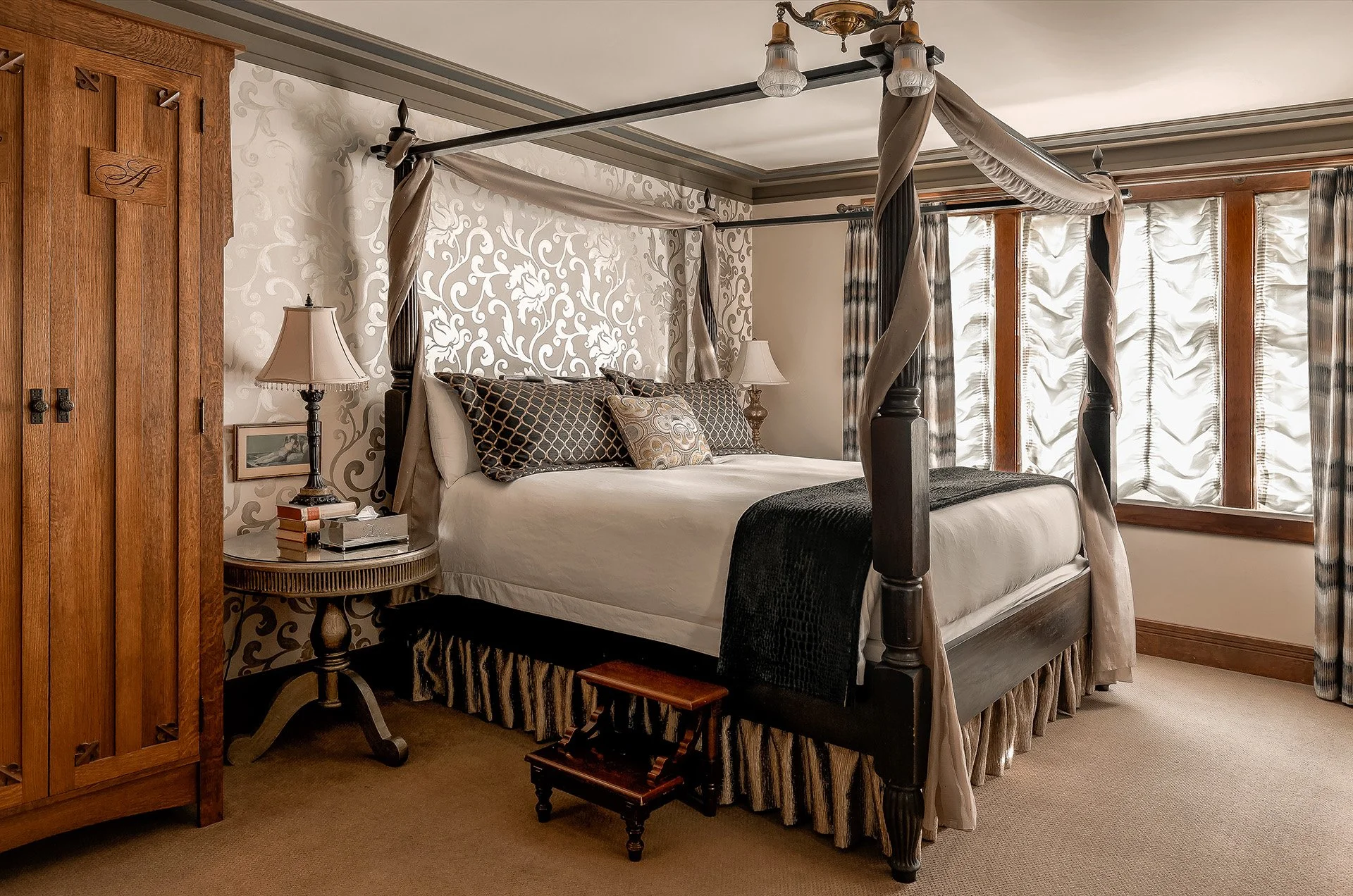A bedroom with a four-poster bed, decorative pillows, a bedside table with lamps, a wooden wardrobe, and large windows with patterned curtains.