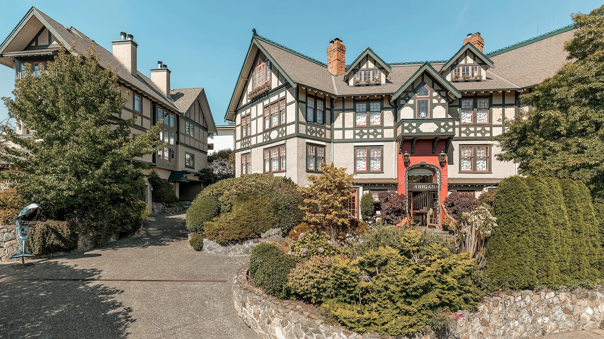 A large, multi-story house with Tudor-style architecture, featuring a mix of stucco and dark wood trim, multiple gabled roofs, and numerous windows. The house is surrounded by well-maintained shrubbery and trees, with a paved driveway leading up to it.