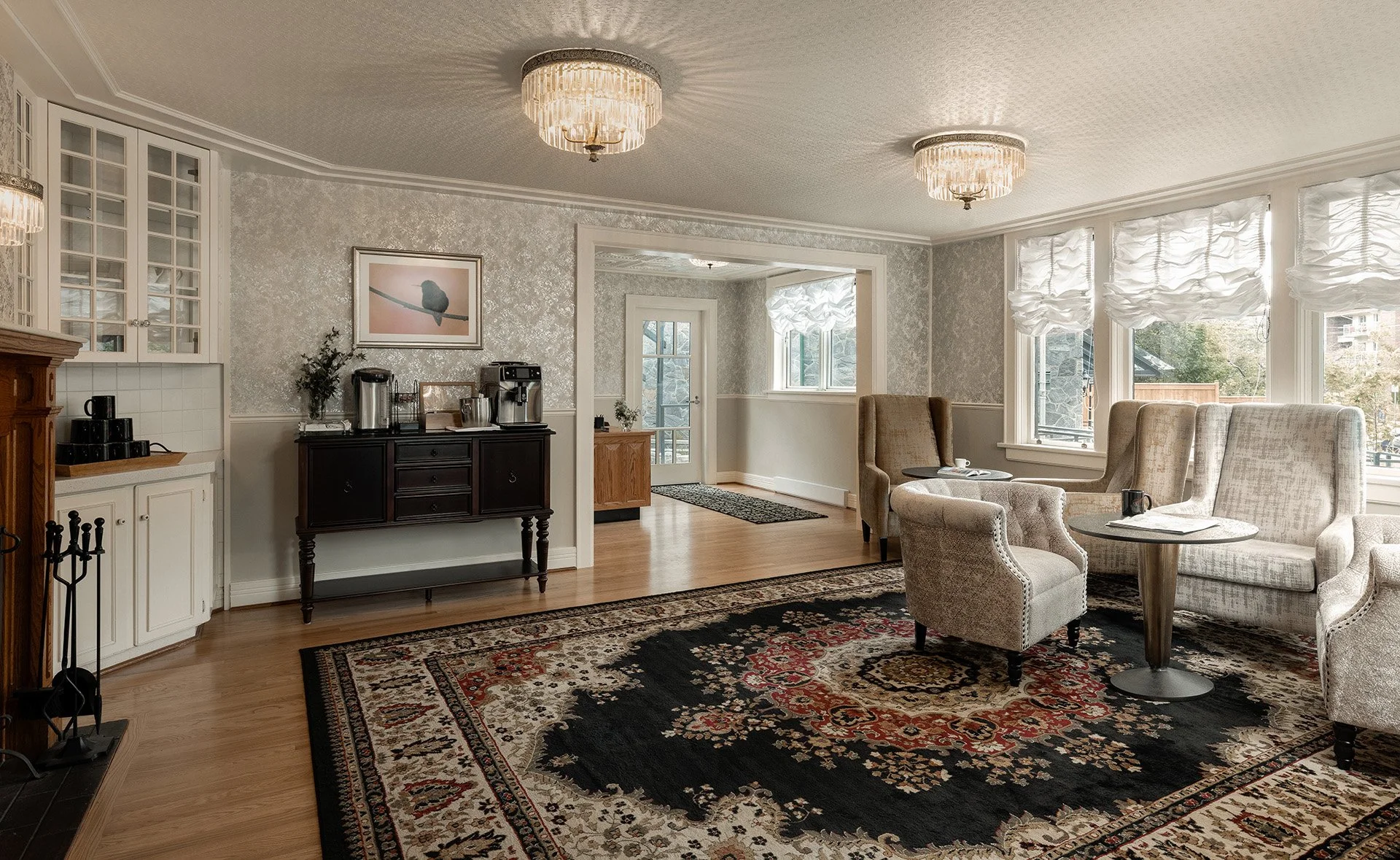 A spacious living room with large windows, patterned wallpaper, a chandelier, a black sideboard with wet coffee equipment, a patterned rug, and chairs and sofas arranged for seating.
