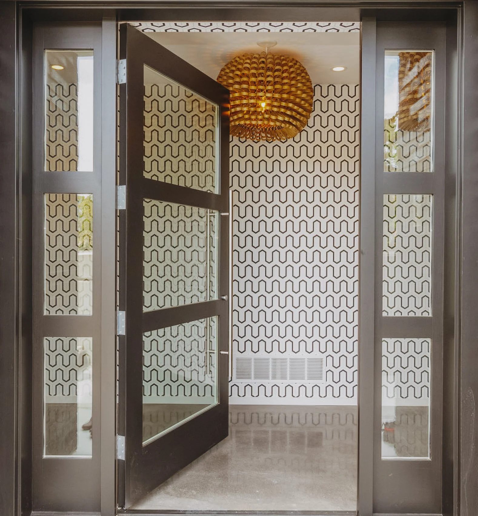 View of an interior possibly an entryway, with open black door, black frame, patterned black and white wallpaper, a wooden pendant light, and a vent on the wall.