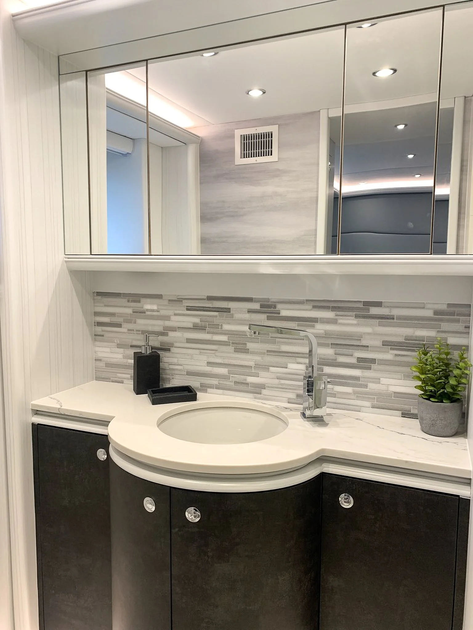 Modern bathroom vanity with a round sink, black cabinetry, a white marble countertop, a mosaic tile backsplash, a small plant in a gray pot, and a mirror with storage behind.