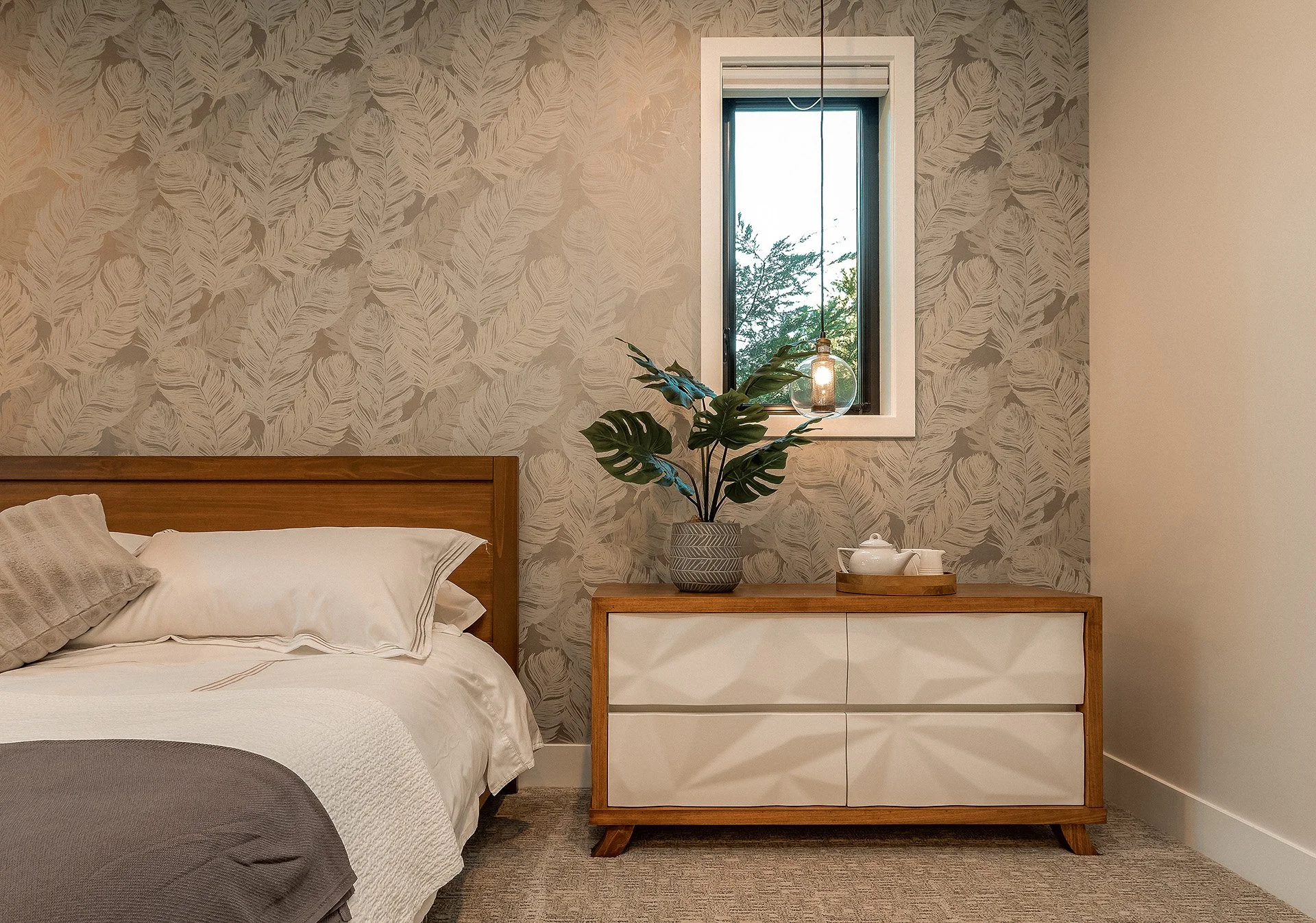 A bedroom corner with a wooden bed, white bedding, gray blanket, and pillows, a wooden and white dresser, a large potted plant on top, a small tray with a tea set, and a window with a view of green trees in the background.