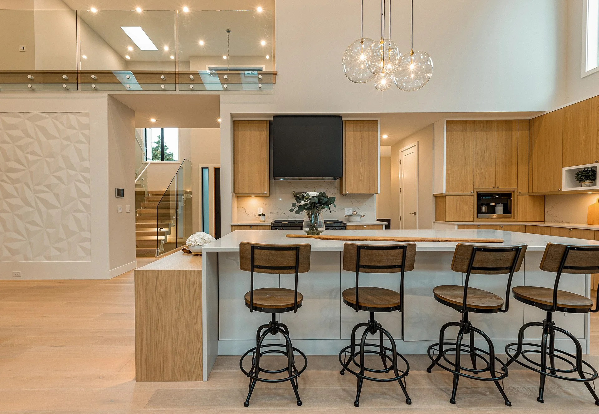 Modern kitchen with wooden cabinets, a white island with four wooden bar stools, a vase with flowers, and a hanging glass light fixture; stairs and a balcony are visible in the background.