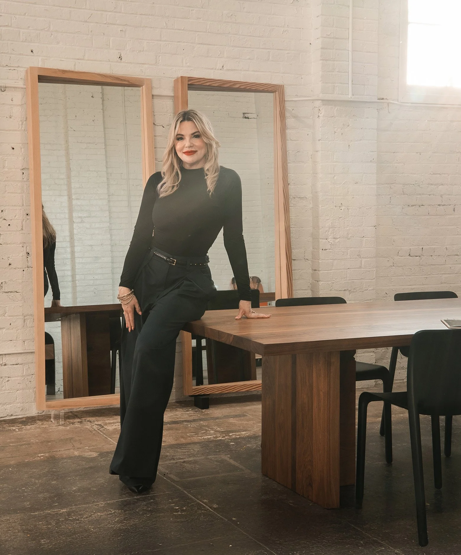 Danisha, a woman with blonde hair, dressed in black, poses in a modern, minimalist room with a brick wall and a large mirror, standing with one knee on a wooden table.