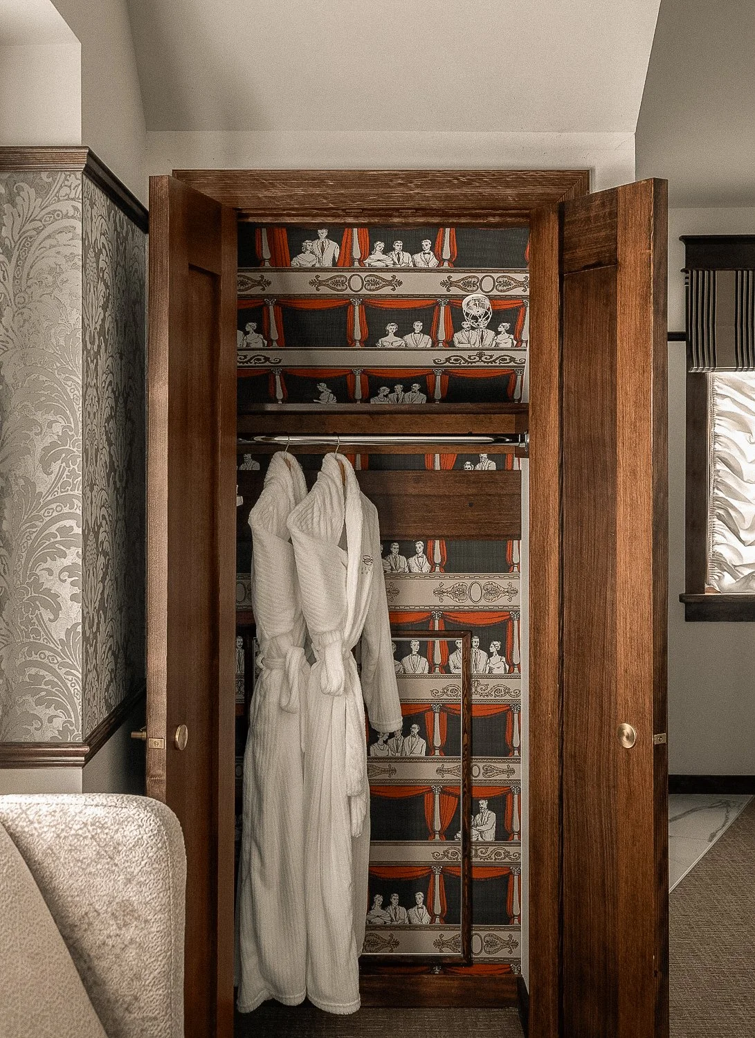 An open wooden closet door reveals a vintage wallpapered interior with theater or circus performers, with two white robes hanging inside.