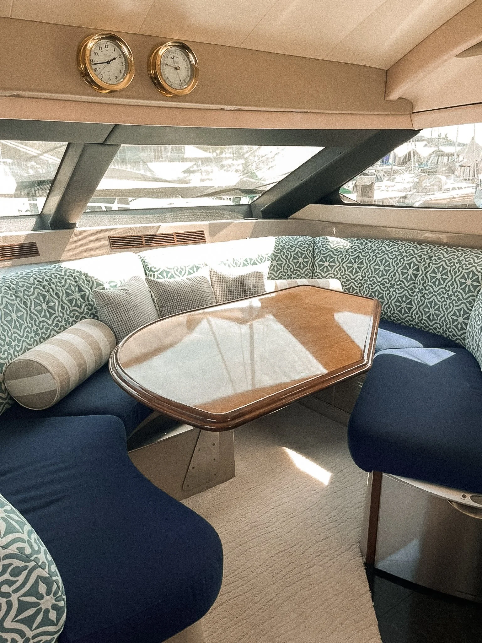 Interior of a boat with a curved sitting area and a wooden table, nautical decor, large window view of boats and marina outside, two clocks on the wall