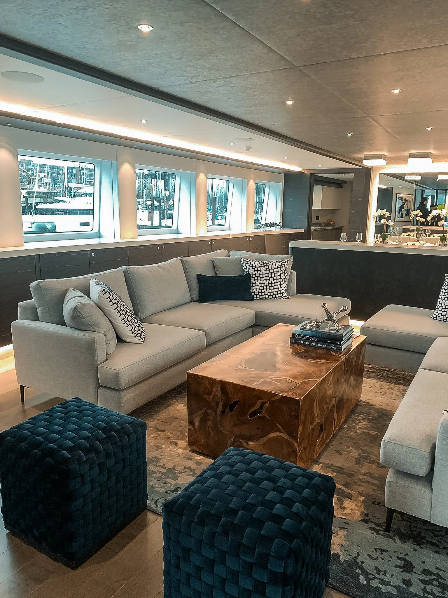 Modern living room with light gray sofas, black and white patterned pillows, a wooden coffee table, and dark blue ottomans, with large windows overlooking a marina.
