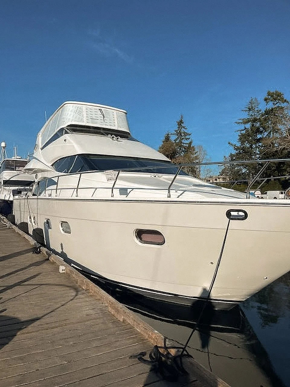White yacht docked at a marina on a sunny day with trees and parked cars in the background.