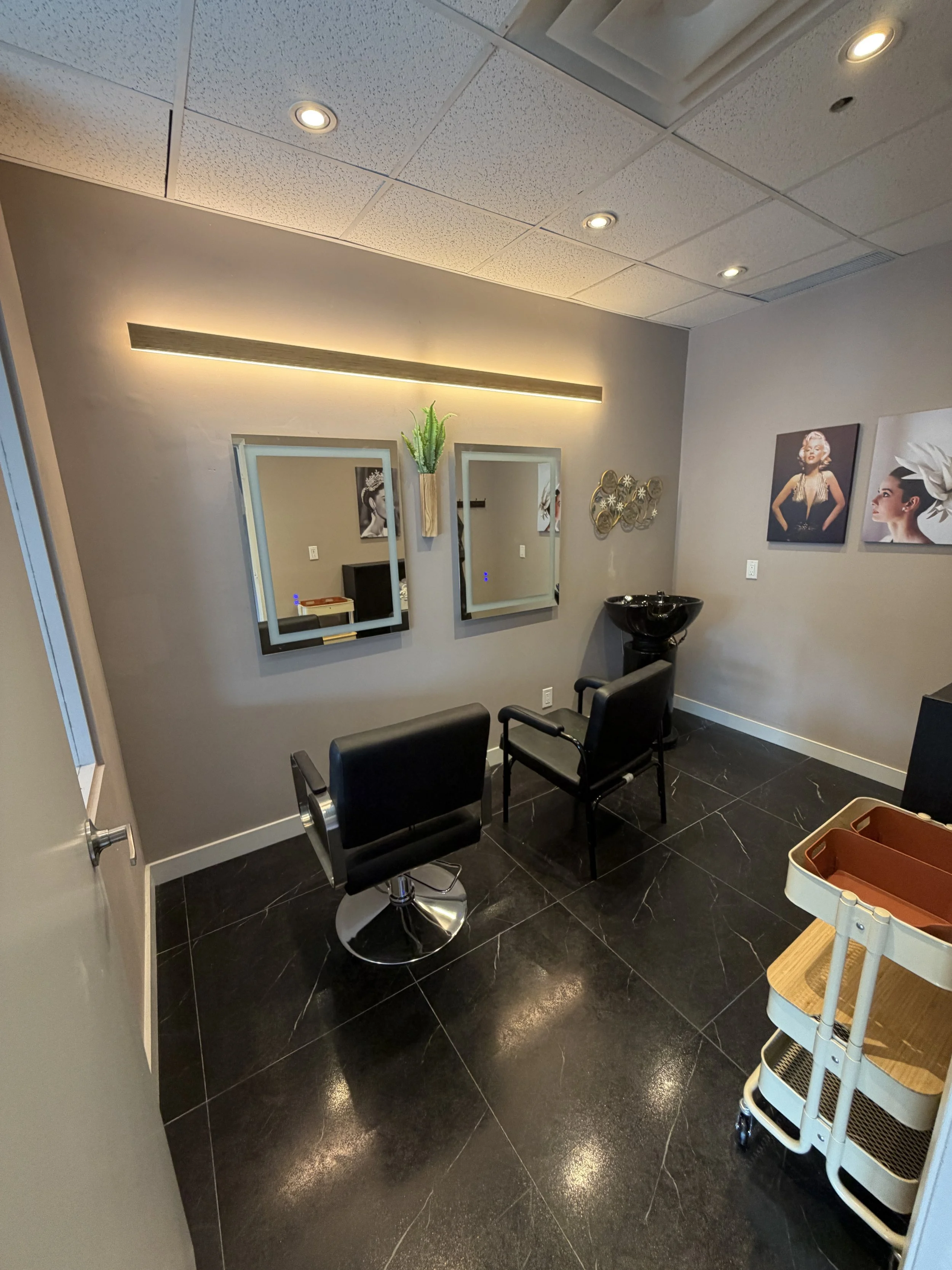 View of a salon or barber shop with two black barber chairs, mirrors on the wall, framed artwork of Marilyn Monroe and a woman with a hat, a black basin for washing hair, and a rolling cart with wooden shelves and baskets on black tiled floor ceiling