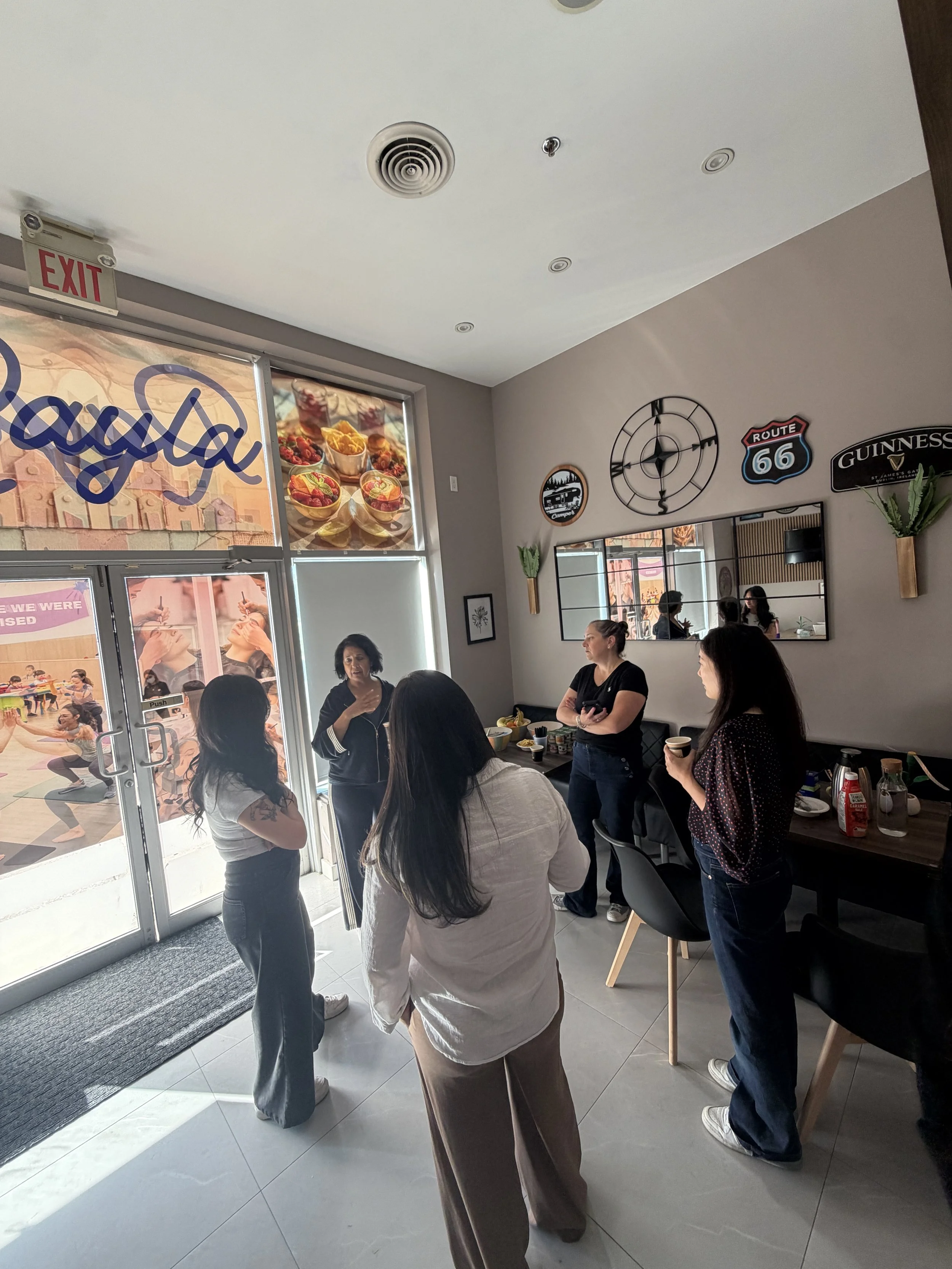 A group of people, mostly women, are gathered inside a restaurant near the entrance, engaged in conversation. Some are holding cups and snacks, and there is a counter with condiments. The restaurant has a window with signs and photos, and decorations