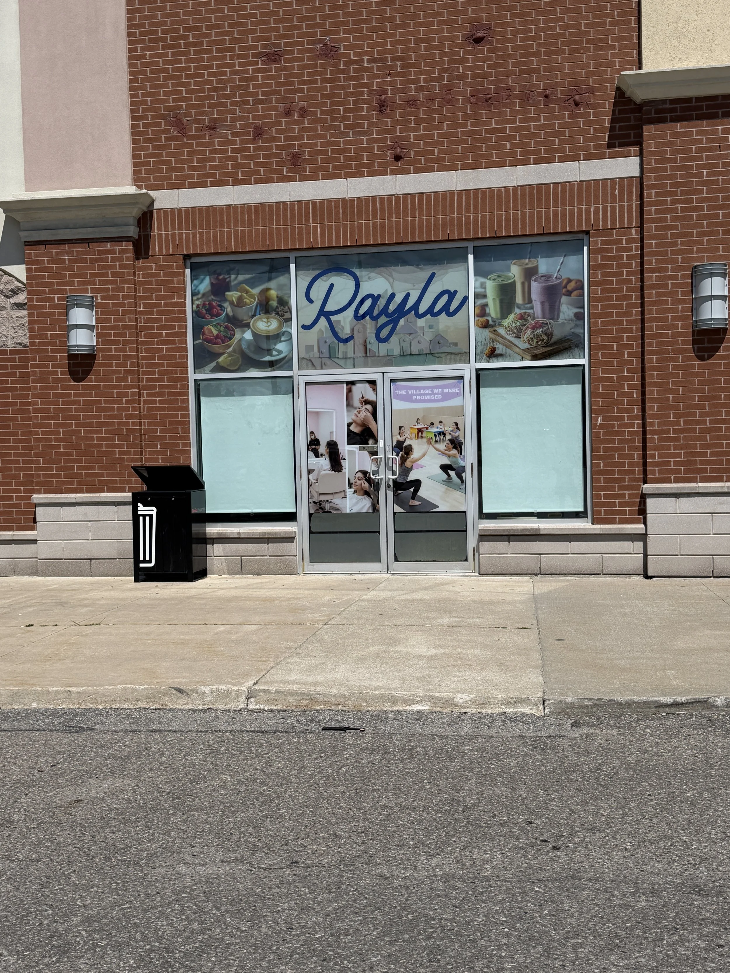 Front view of a brick storefront with large glass windows displaying images of food items and the name 'Rayla' in blue cursive letters above the door. There are two modern outdoor wall sconces on either side of the storefront, a black trash bin to the left, and a concrete sidewalk in front.