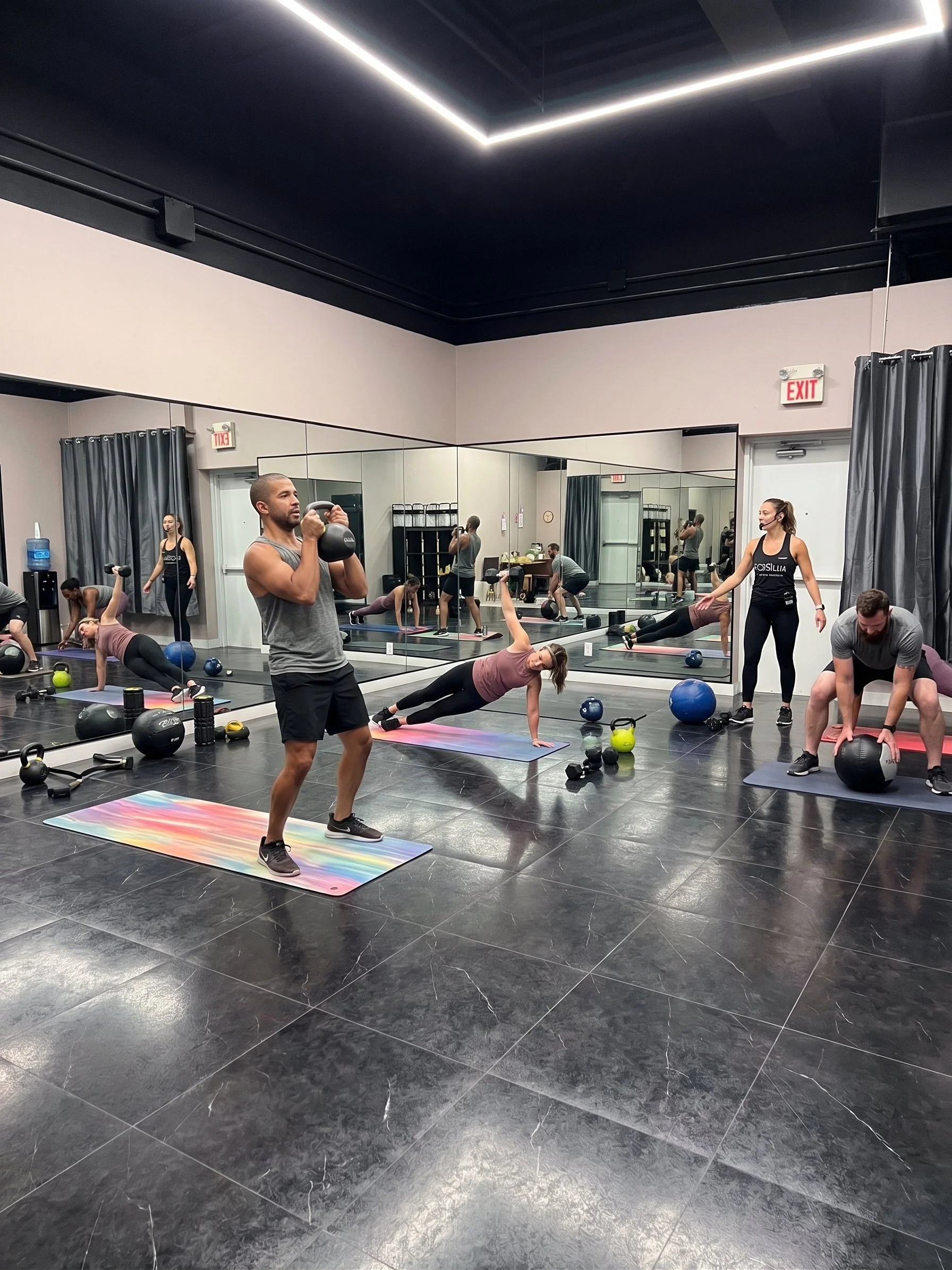 People participating in a fitness class in a gym with black tiled floor and a large mirror wall, some using exercise balls and kettlebells, a yoga mat, and a fitness instructor leading the session.