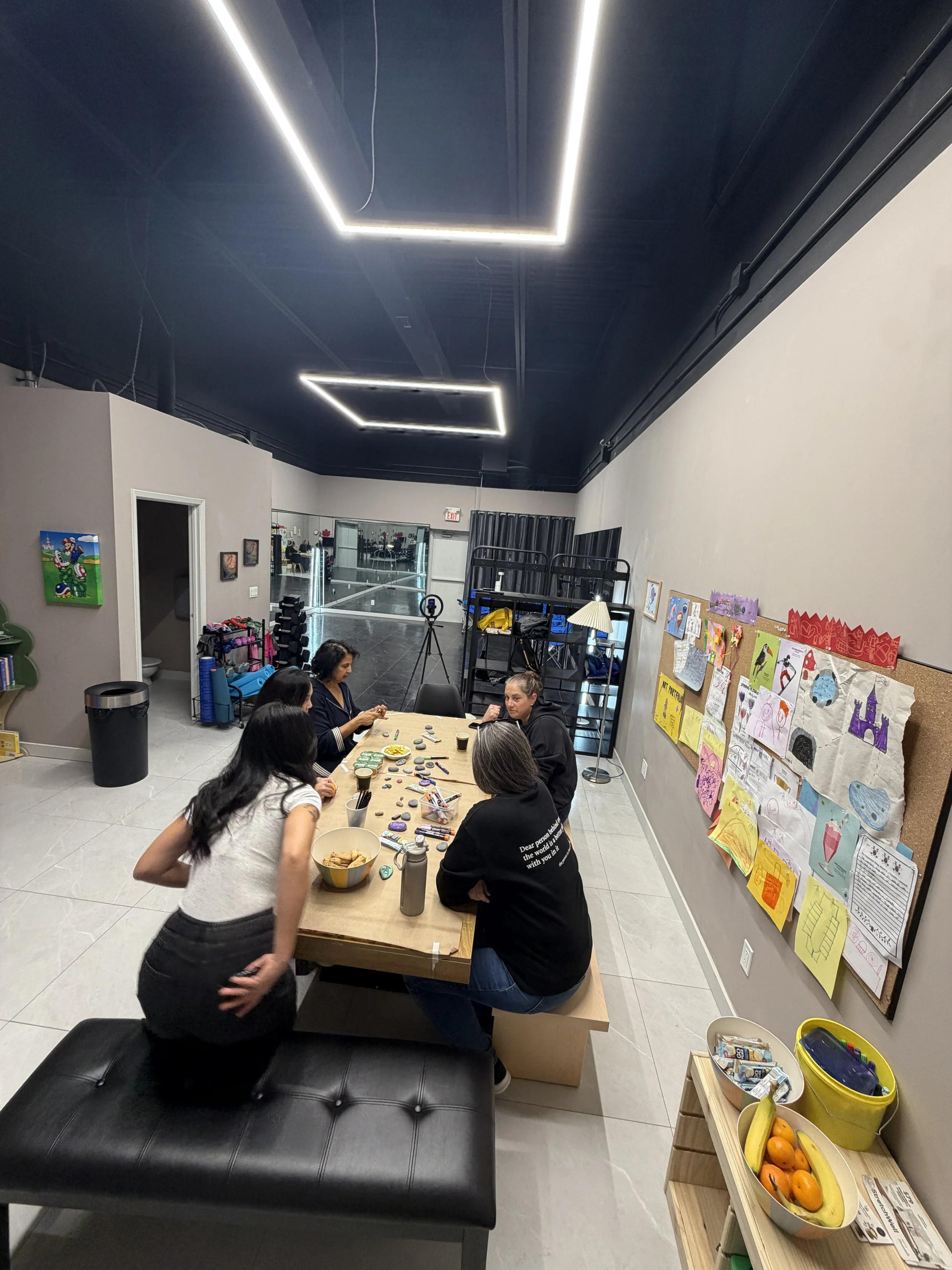People gathered around a table with arts and crafts materials in an indoor room with artwork on the walls and a bulletin board decorated for a holiday.