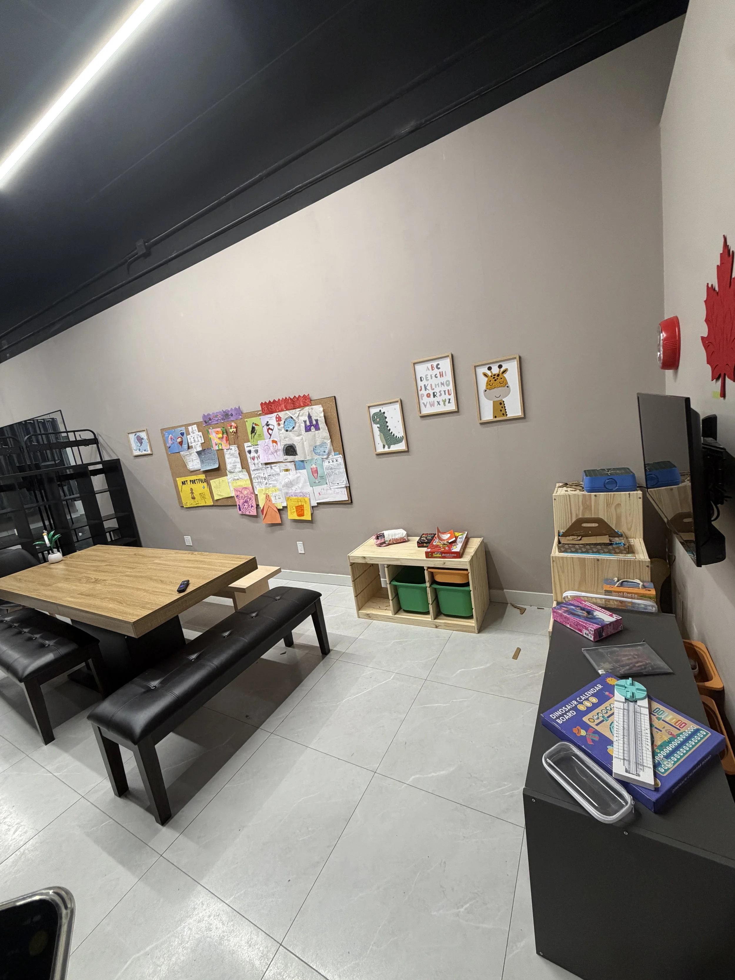 A modern classroom or office space with a large wooden table and black cushioned benches. The wall features children's artwork and decorations, including framed animal illustrations and a colorful bulletin board. A small black table holds educational materials and a remote, with a black shelving unit and a cardboard box containing supplies nearby. The floor is tiled with light gray tiles.