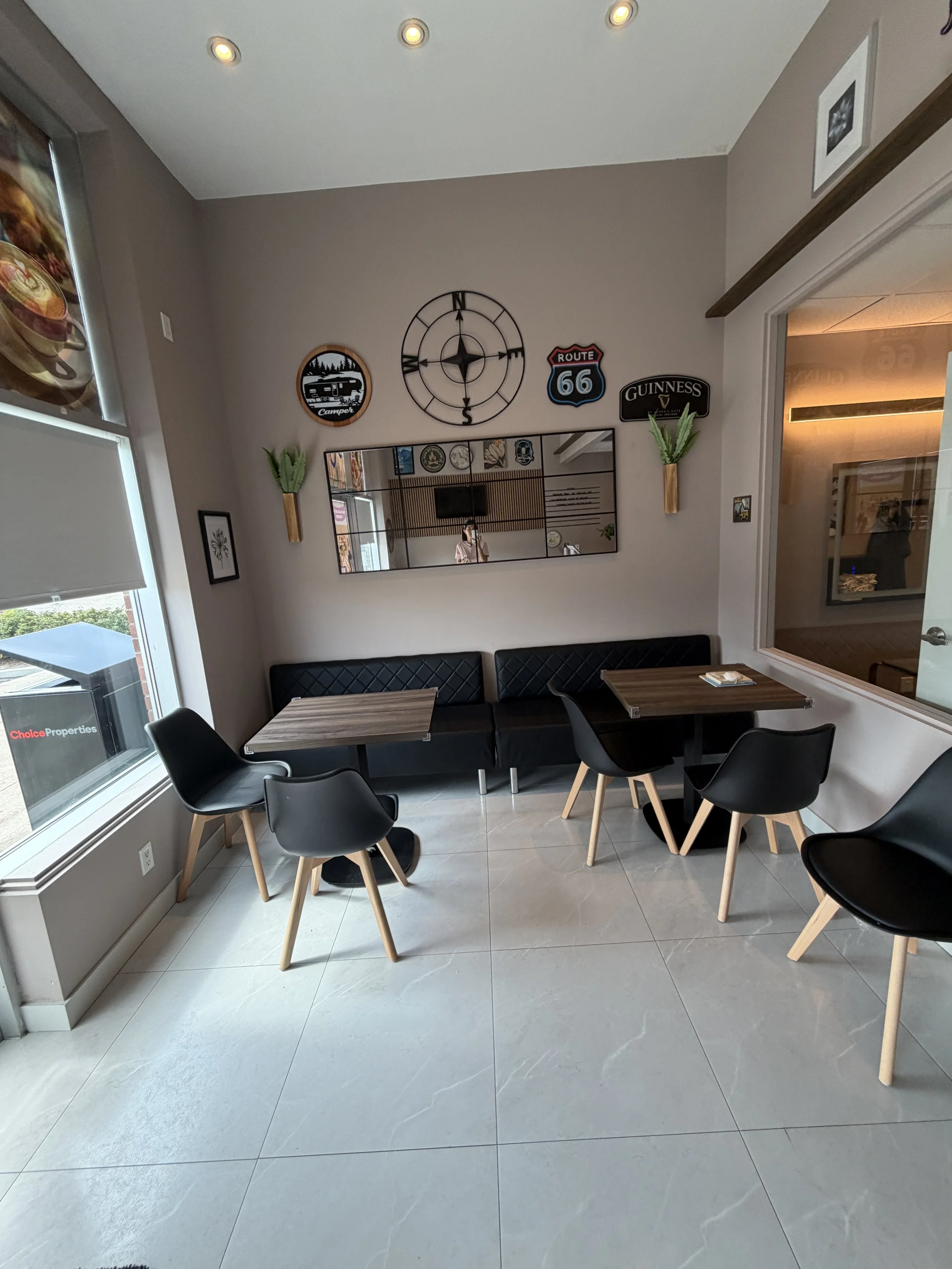 Indoor corner with modern decor, black chairs around small square tables, gray tiled floor, wall decor including a large clock, Route 66 sign, Guinness sign, small plants, and a mirror reflecting the room.