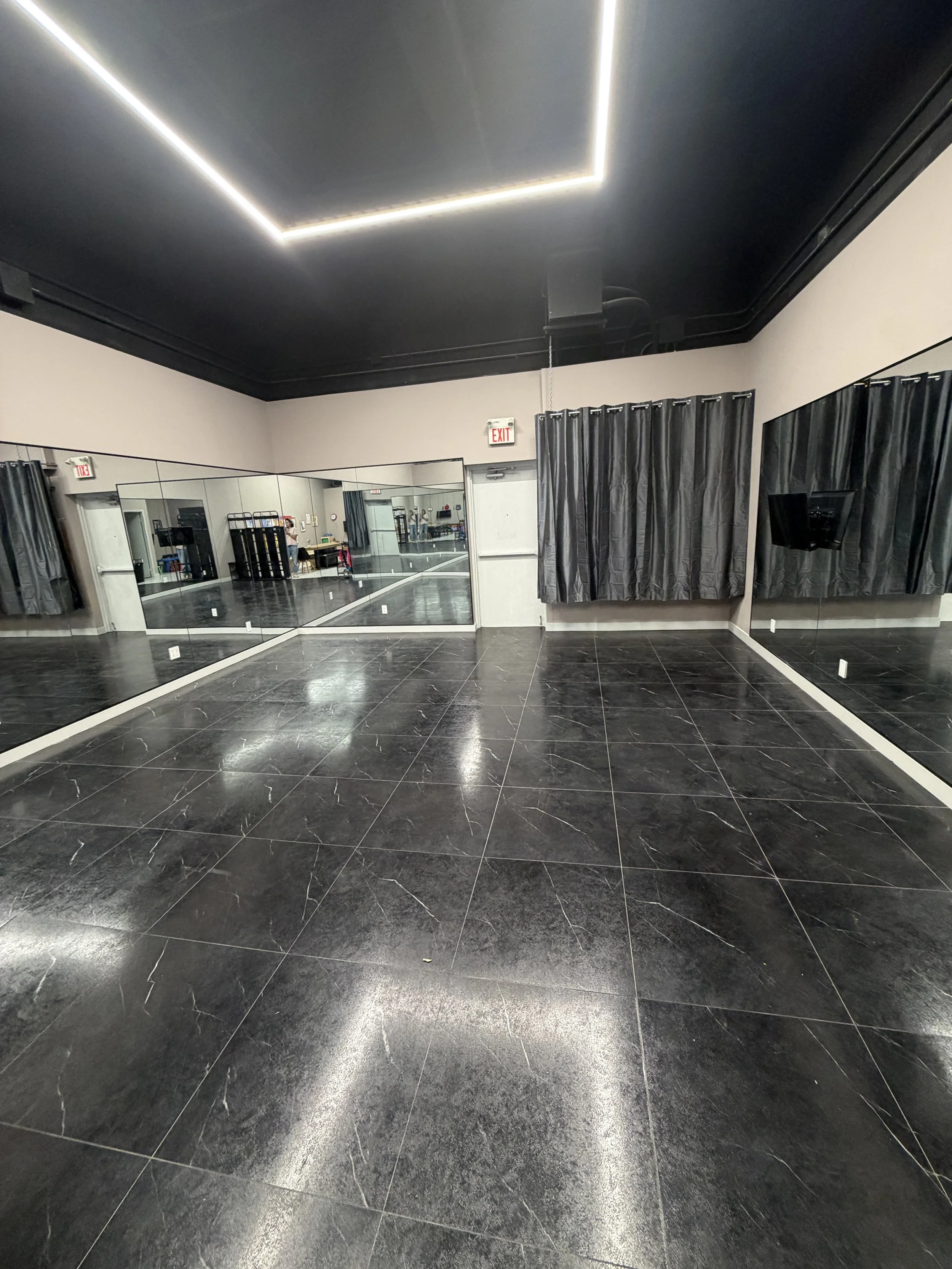 An empty dance studio with black tiled floor, large wall mirrors, black curtains, and a ceiling with black paint and LED strip lighting.