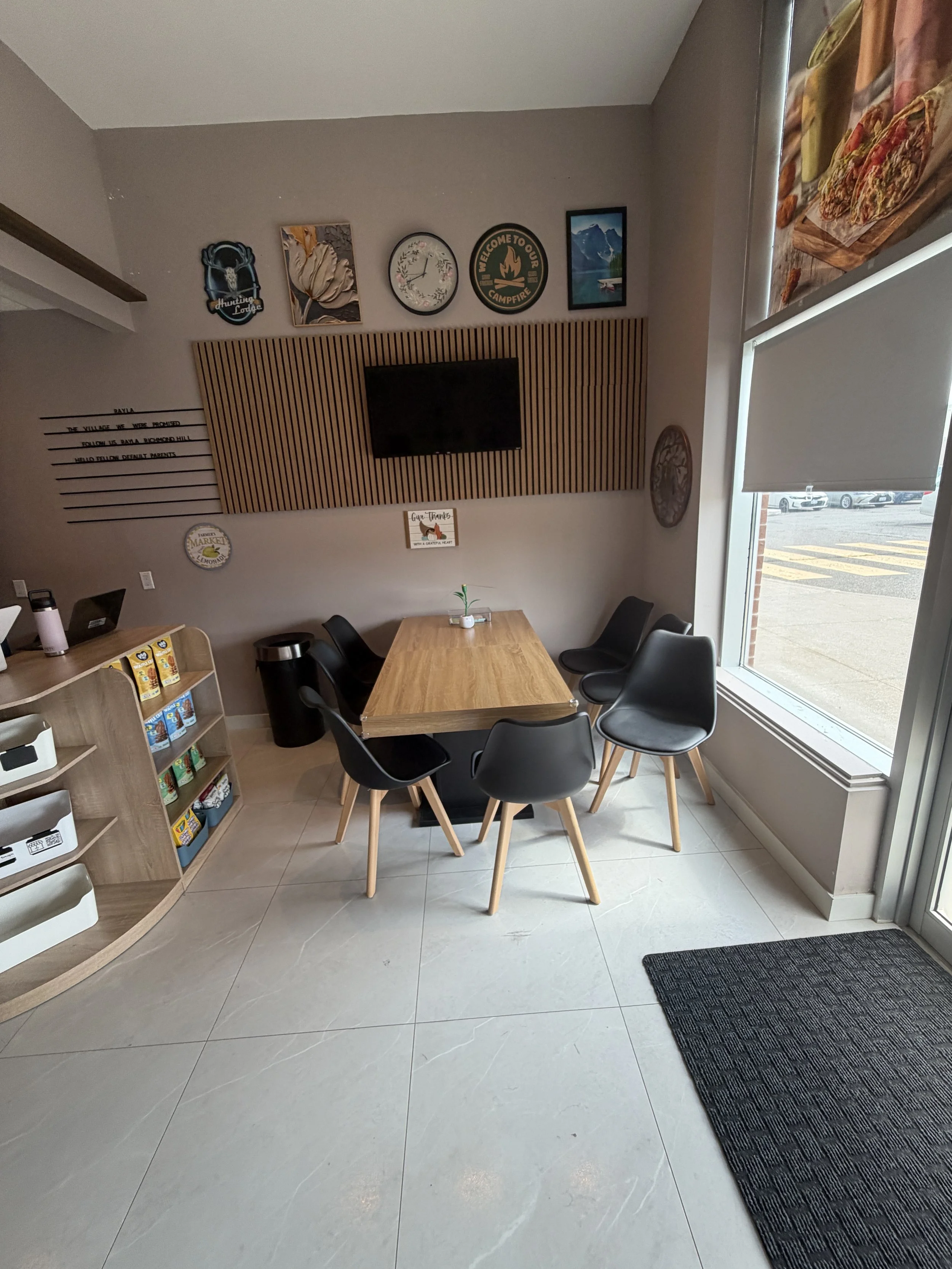 A cozy cafe or restaurant interior with a wooden table and black chairs, a wall decorated with picture frames and signs, and a large window with blinds letting in natural light.