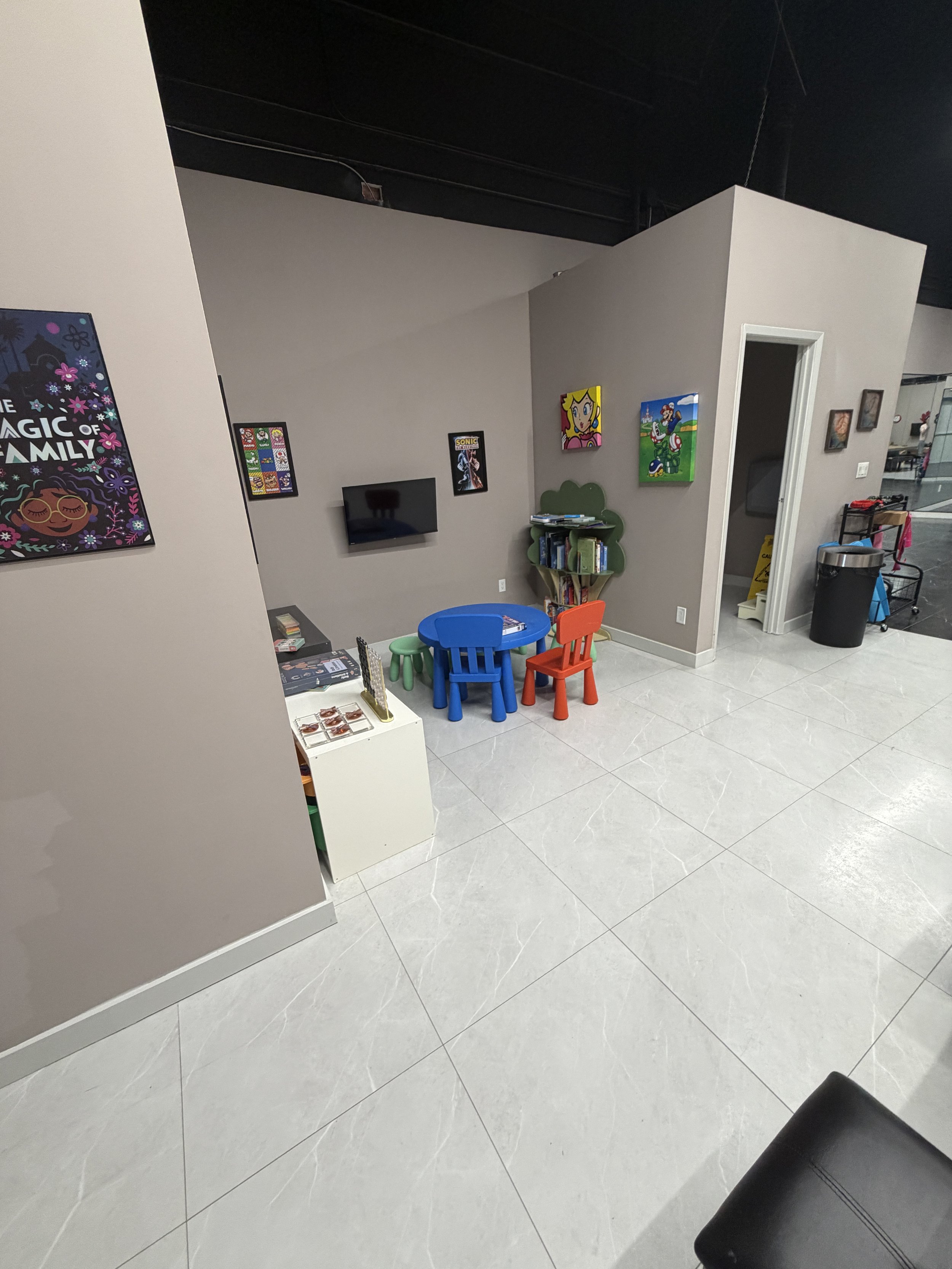 A children's play area with a small blue round table, colorful chairs, a bookshelf with books, and various wall decorations including cartoon characters and framed artwork. There's also a trash can and cleaning supplies nearby.