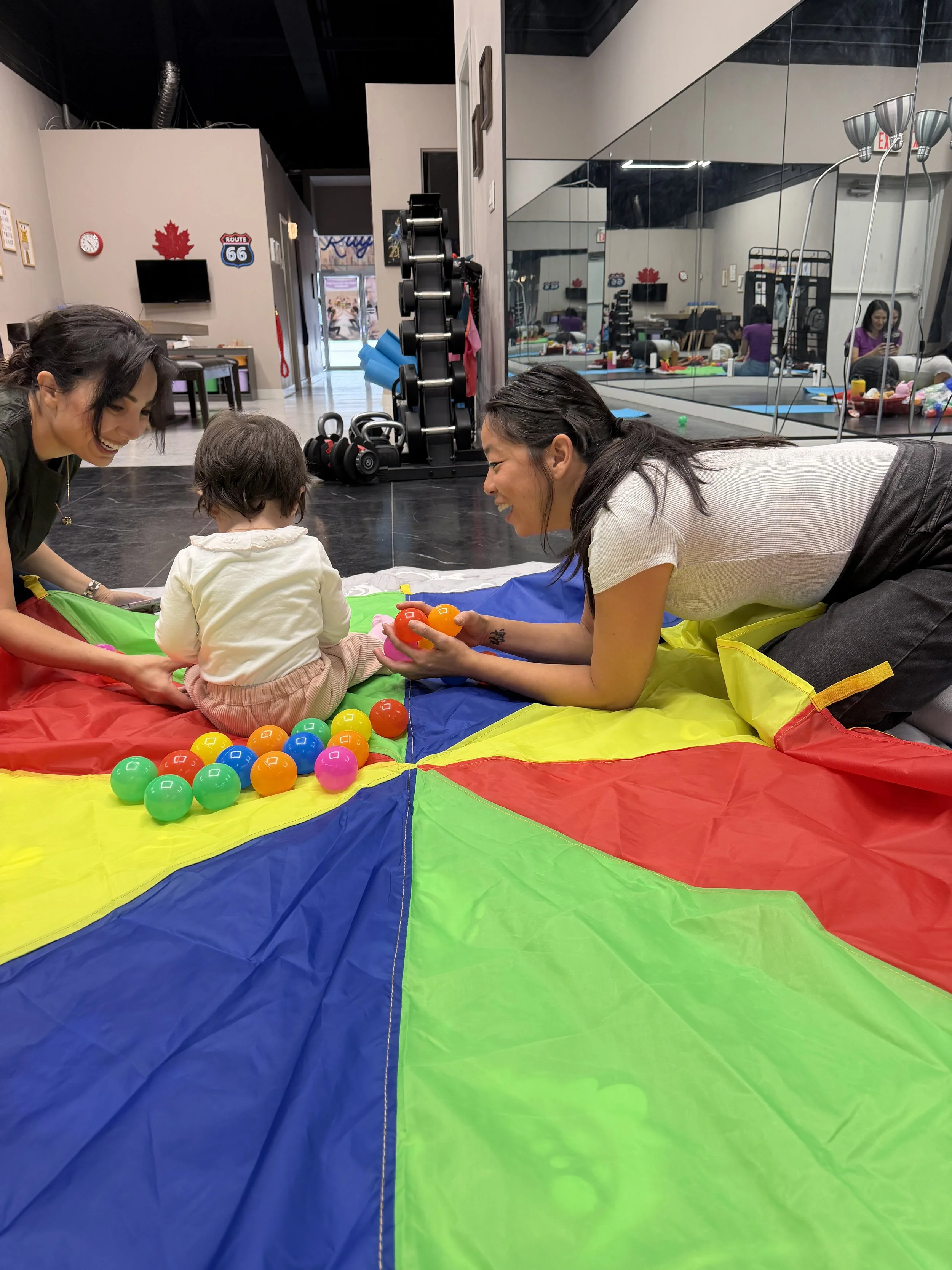 Two women and a small child playing on a colorful mat with plastic balls in an indoor gym or play area, with gym equipment and a mirror on the walls.