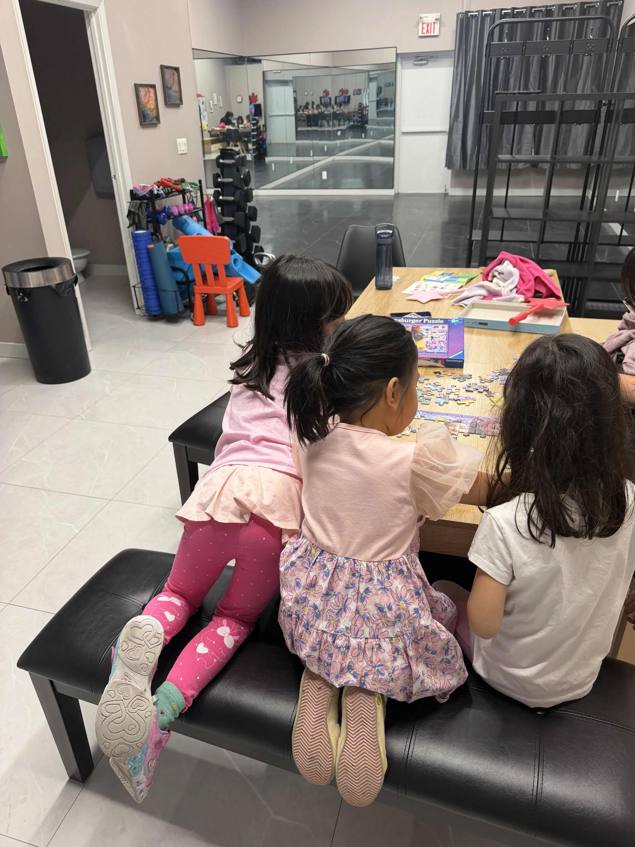 Three children are sitting on a bench and leaning forward, with their legs hanging down, in a room with a table. The children are gathered around the table, looking at something out of view.