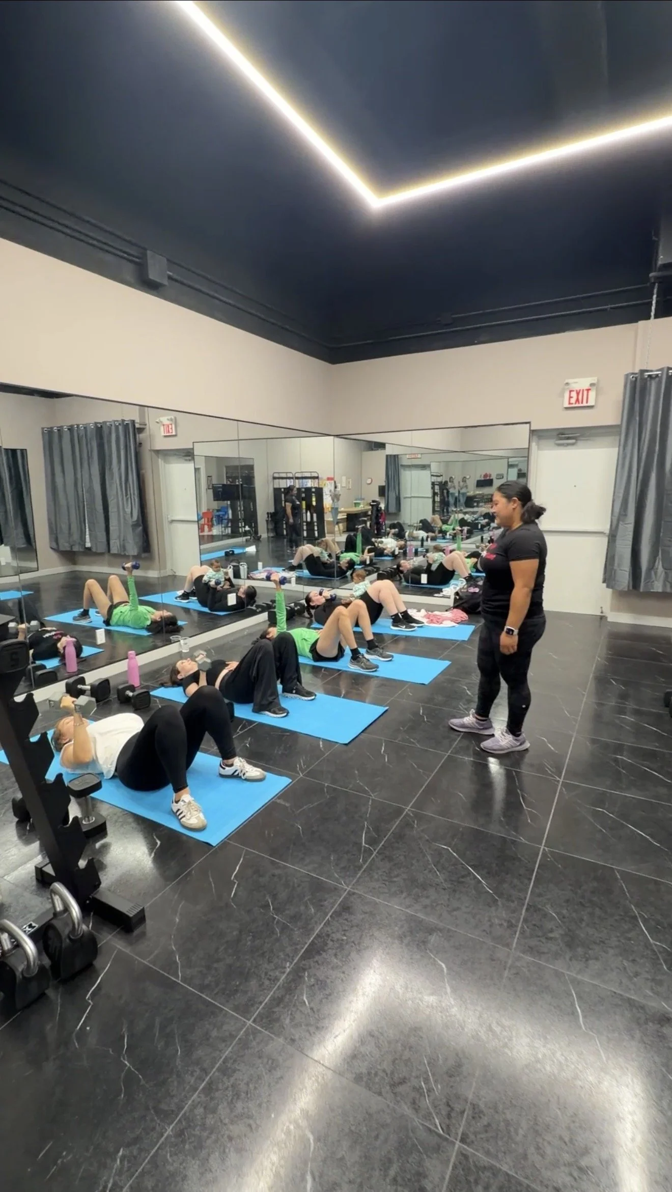 A group fitness class in a gym studio, with women lying on blue mats doing exercises under the instruction of a trainer standing nearby. The room has mirrored walls, black tiled flooring, and LED lighting on the ceiling.