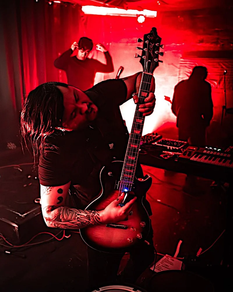 A musician with tattoos on their arm playing an electric guitar on stage during a performance, with two other band members in the background under red stage lighting.
