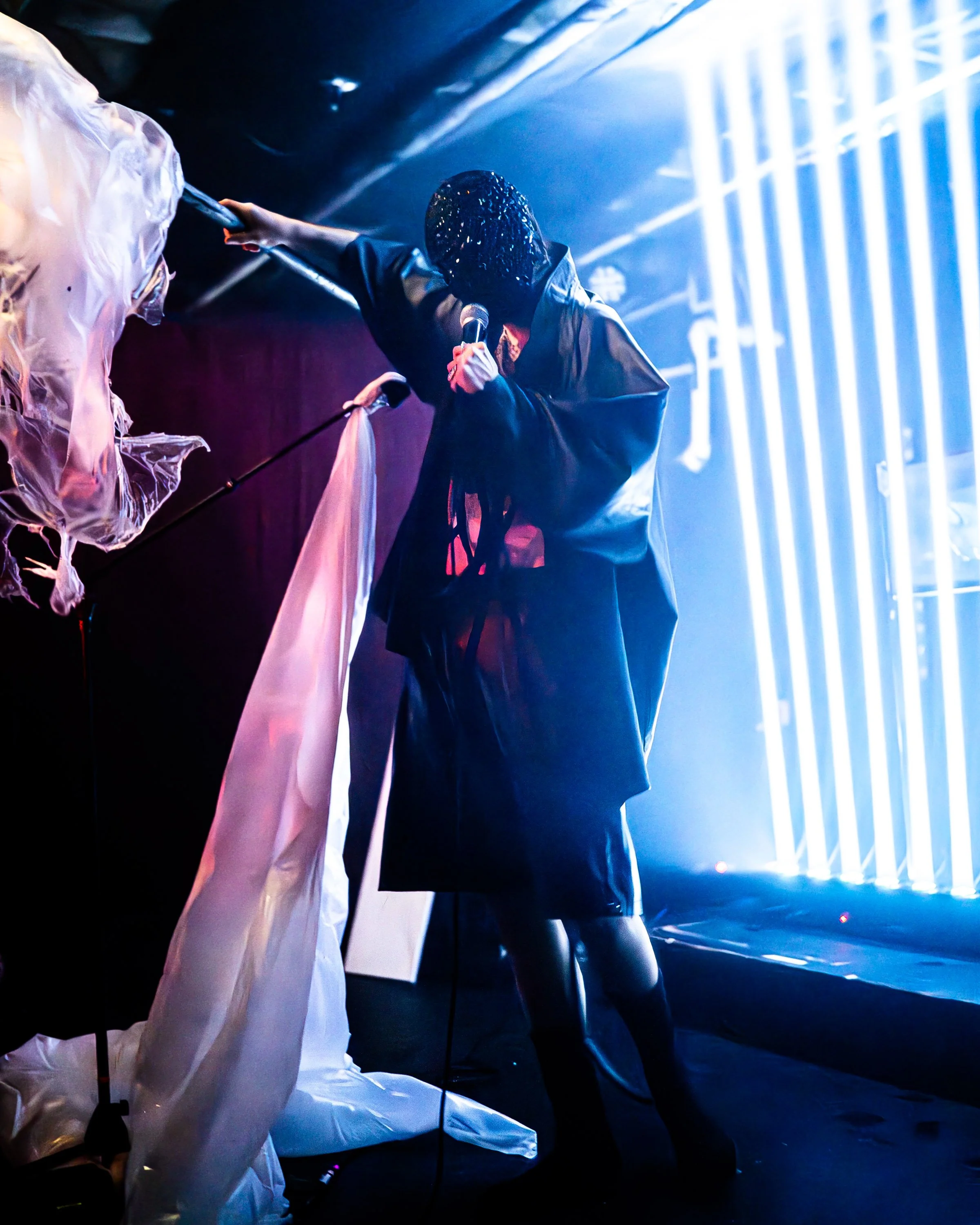A performer on stage, wearing dark clothing and a mask, holding a microphone in one hand and touching a large, flowing fabric with the other, lit by vibrant blue and pink stage lights.