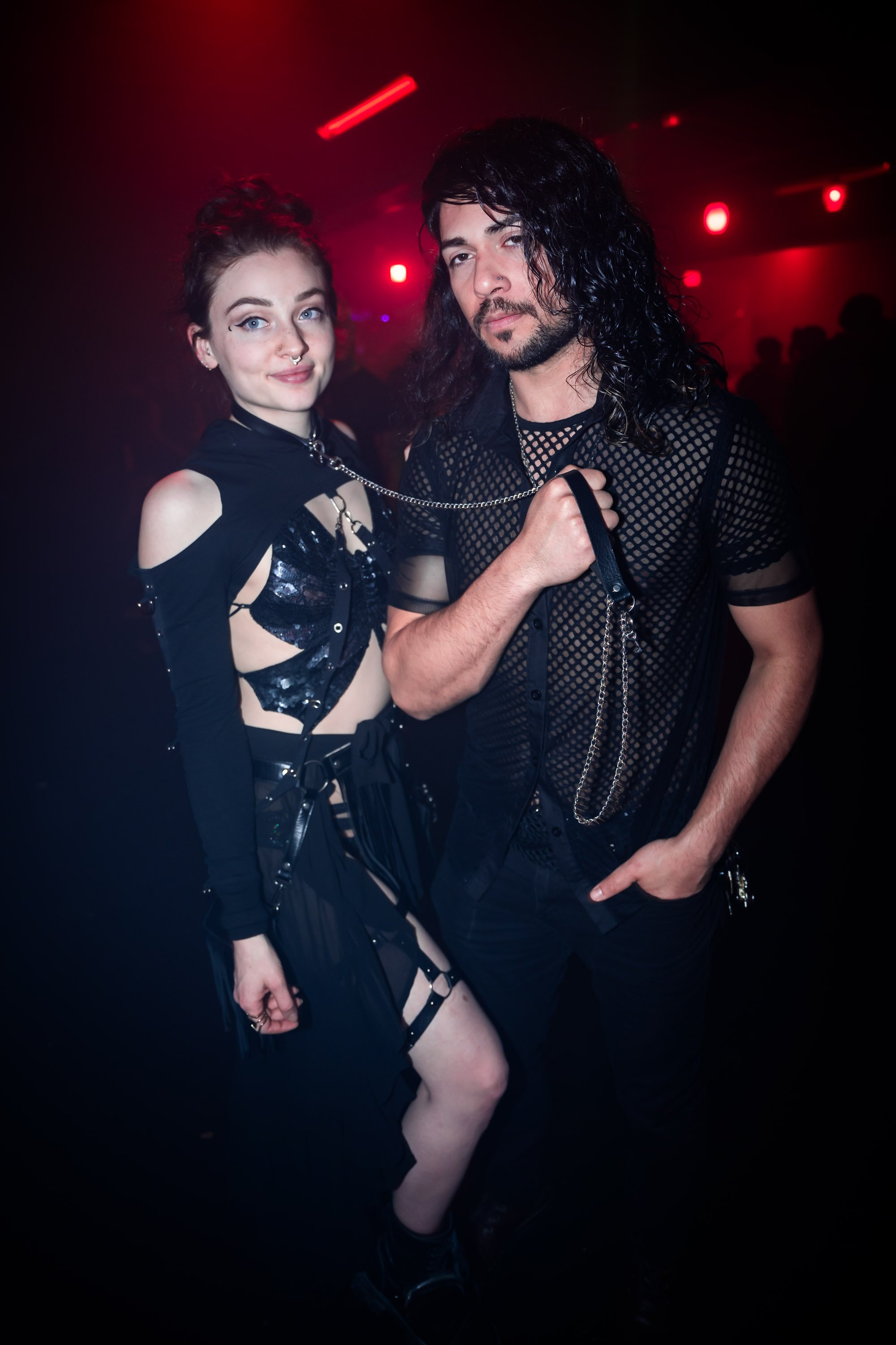 Two individuals dressed in goth or punk attire posing in a dimly lit club with red lights.