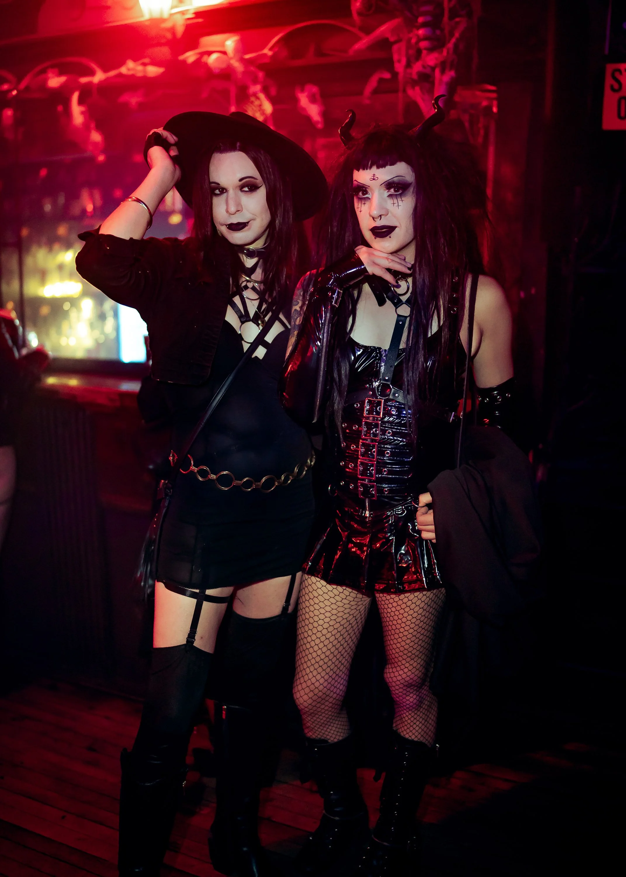 Two women dressed in gothic fashion posing in a dimly lit bar with neon lighting, one wearing a witch hat and the other with devil horns.