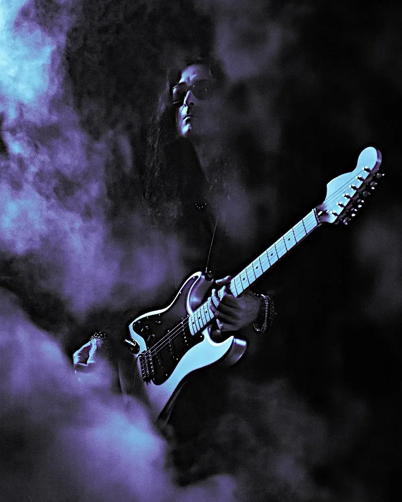 A woman playing an electric guitar in a smoky, dark setting with purple and blue lighting.