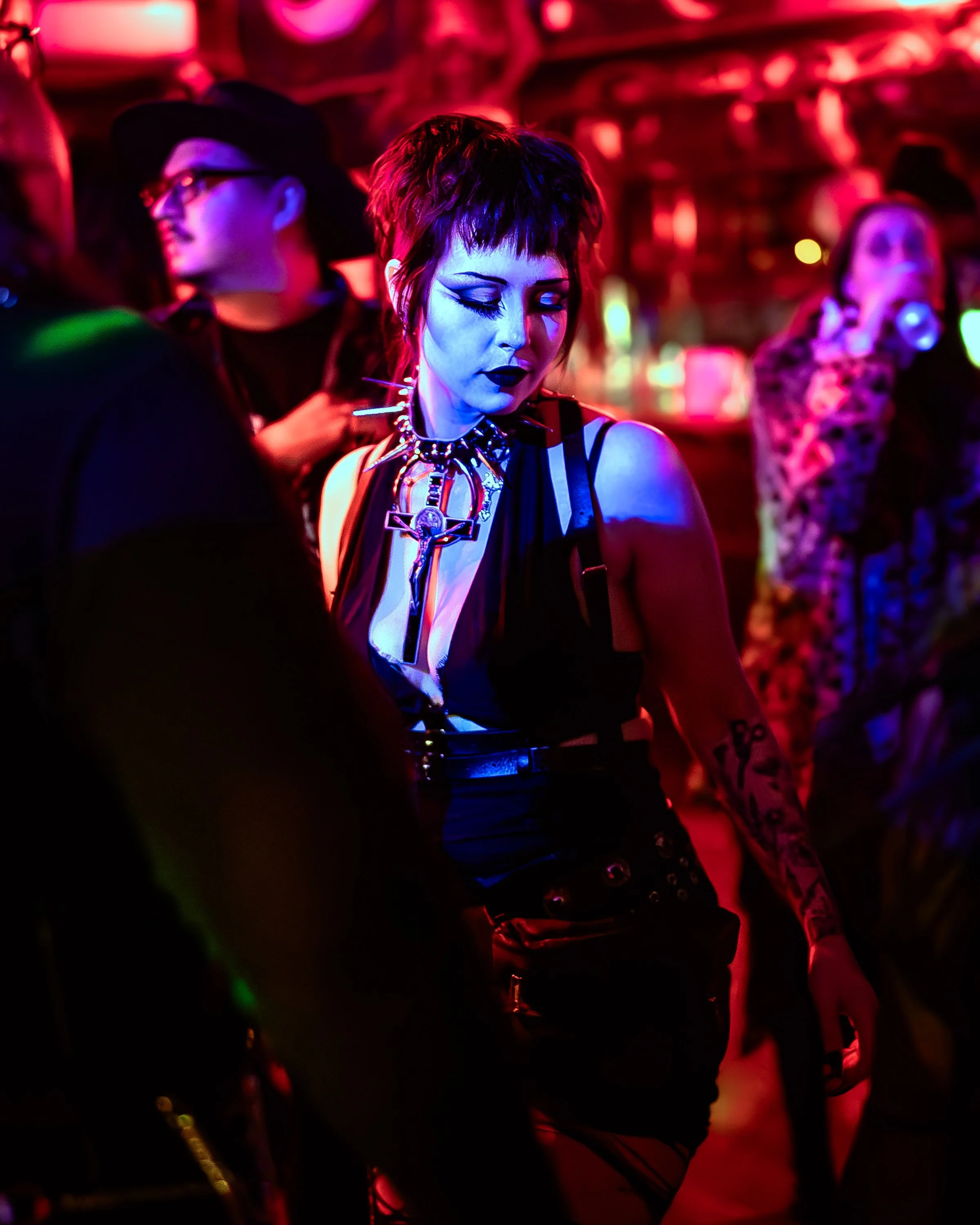 Person with short dark hair and dark makeup, wearing black clothing, jewelry, and tattoos, dancing or socializing in a neon-lit nightclub.