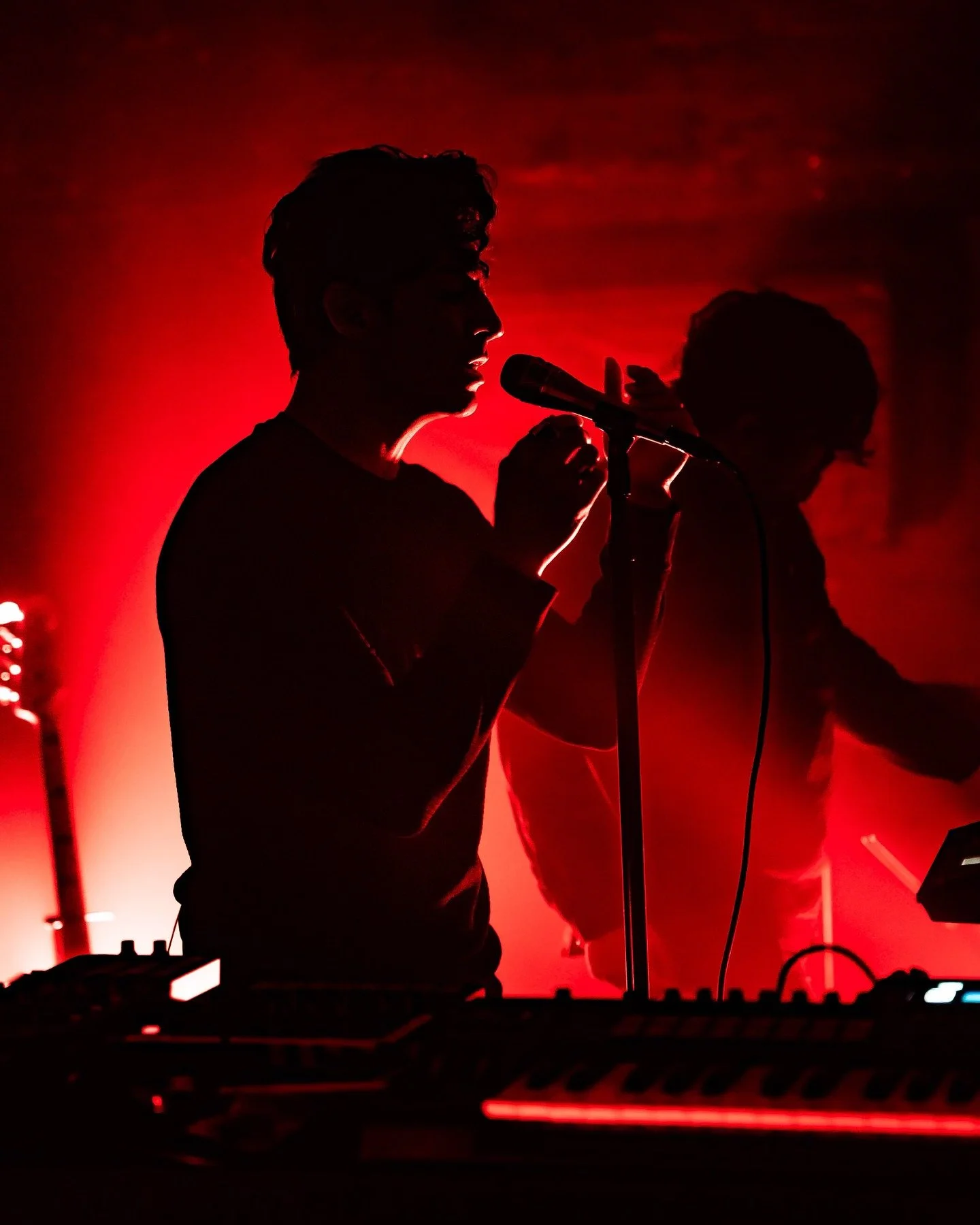 Silhouetted man singing into a microphone on stage with red lighting, accompanied by a second person working electronic equipment.