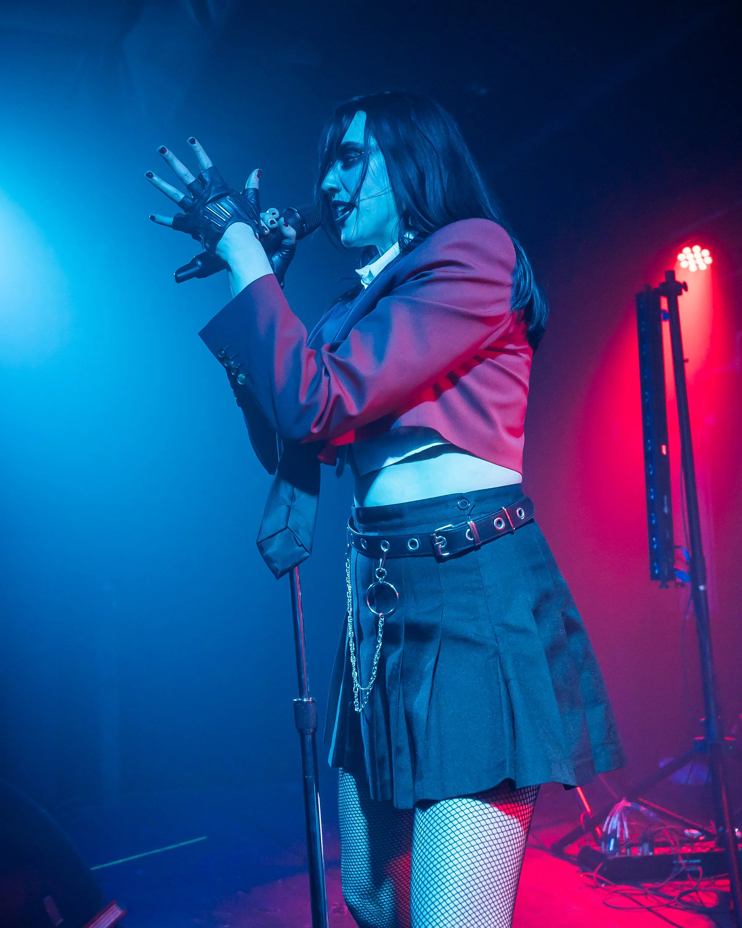 A female singer with dark hair performing on stage, wearing a red blazer, a black pleated skirt, fishnet tights, fingerless gloves, and a belt with chains, illuminated by red and blue stage lights.