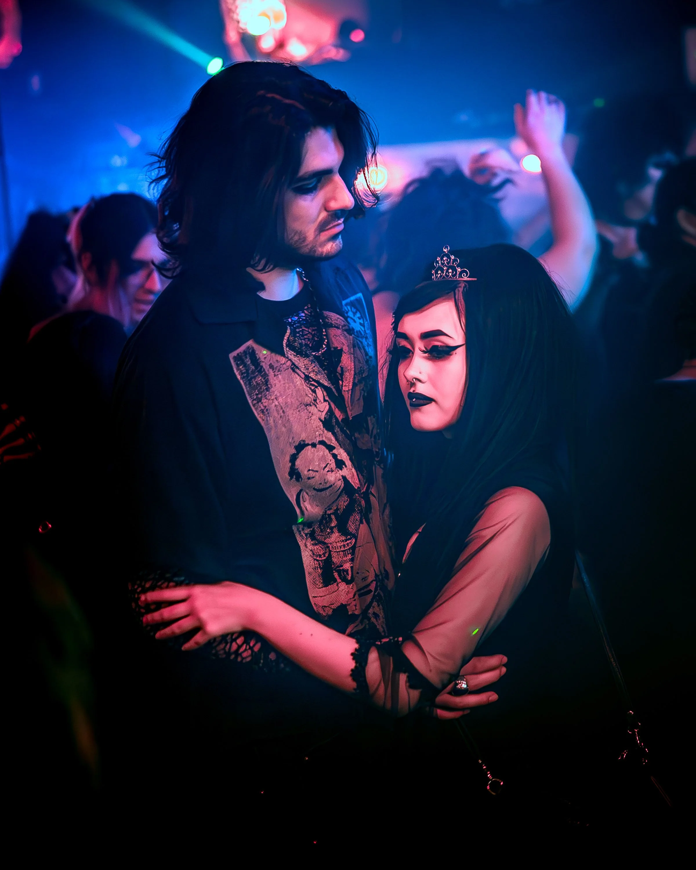 A young couple with gothic style clothing and makeup dancing closely at a nightclub or party, with colorful lights and other people in the background.