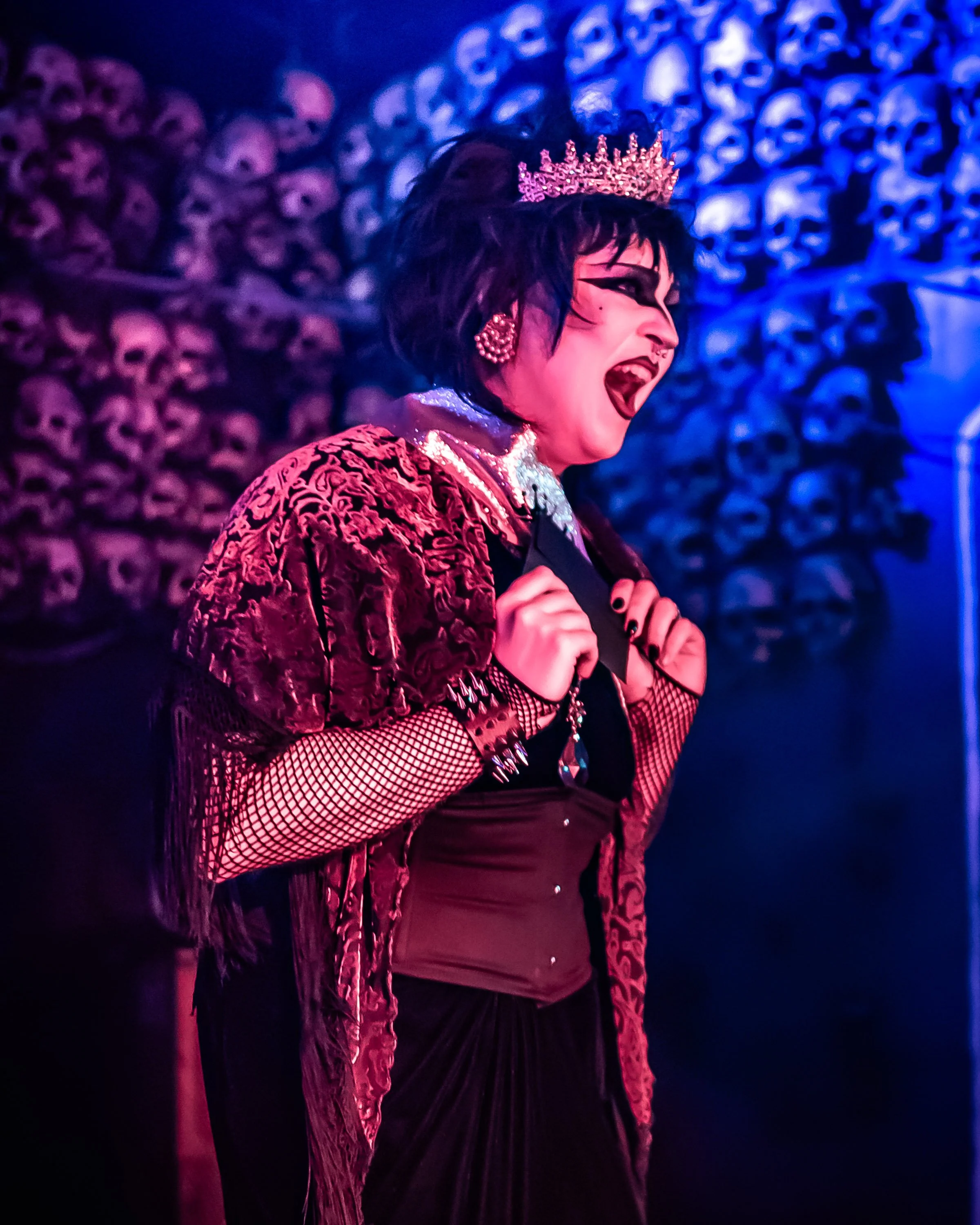 A person dressed in dark gothic style with a crown, black makeup, fishnet gloves, and jewelry, standing in front of a wall of skulls, singing or shouting with a dramatic expression.