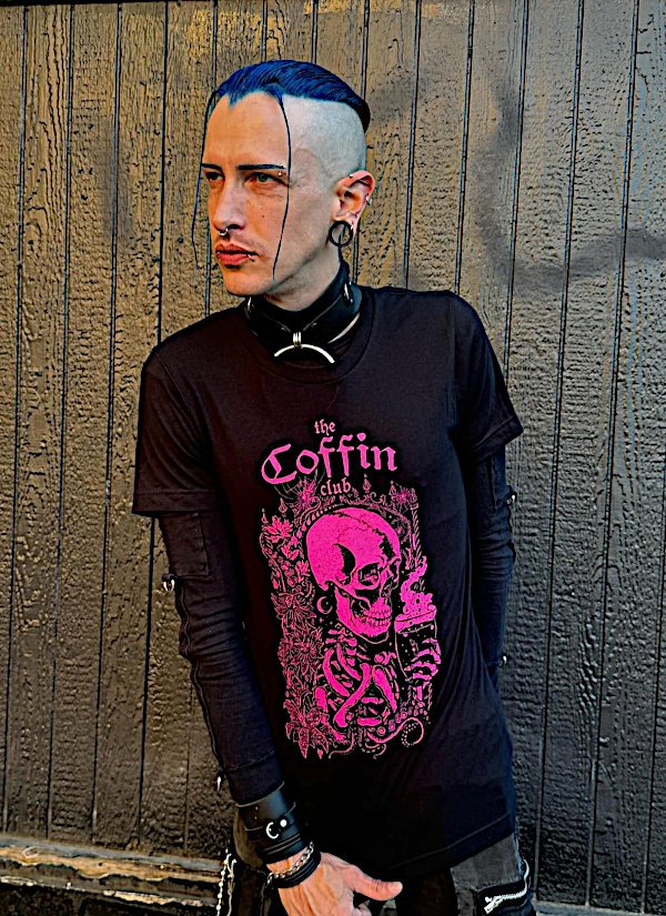 A person with blue hair, partially shaved on one side, wearing black clothing with pink graphic print of a skull and the words 'the coffin club,' standing in front of a wooden fence.