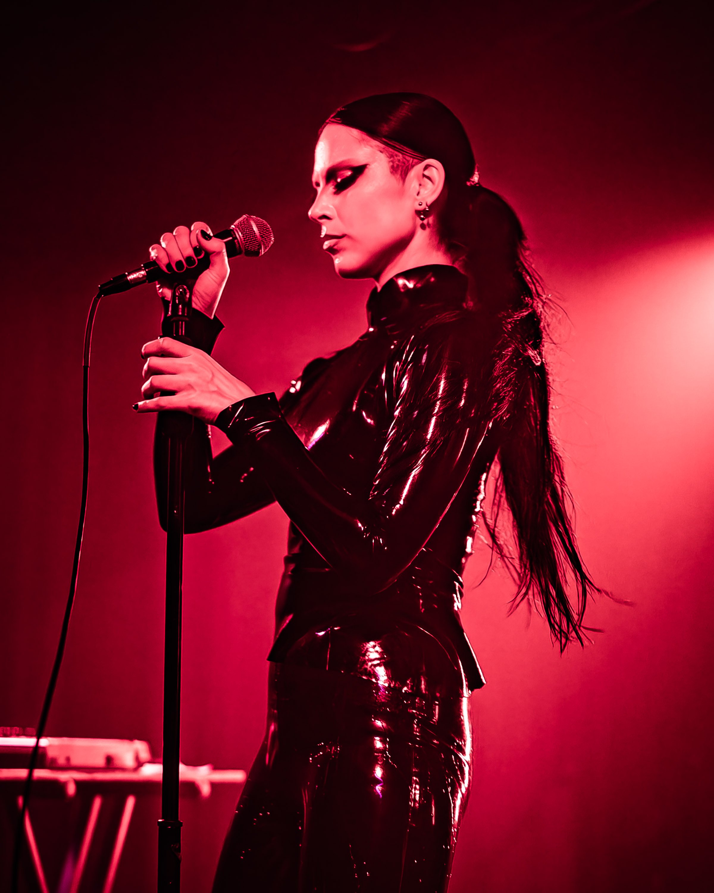 A woman singing into a microphone during a performance, dressed in a shiny black outfit with dark makeup, illuminated by red stage lighting.