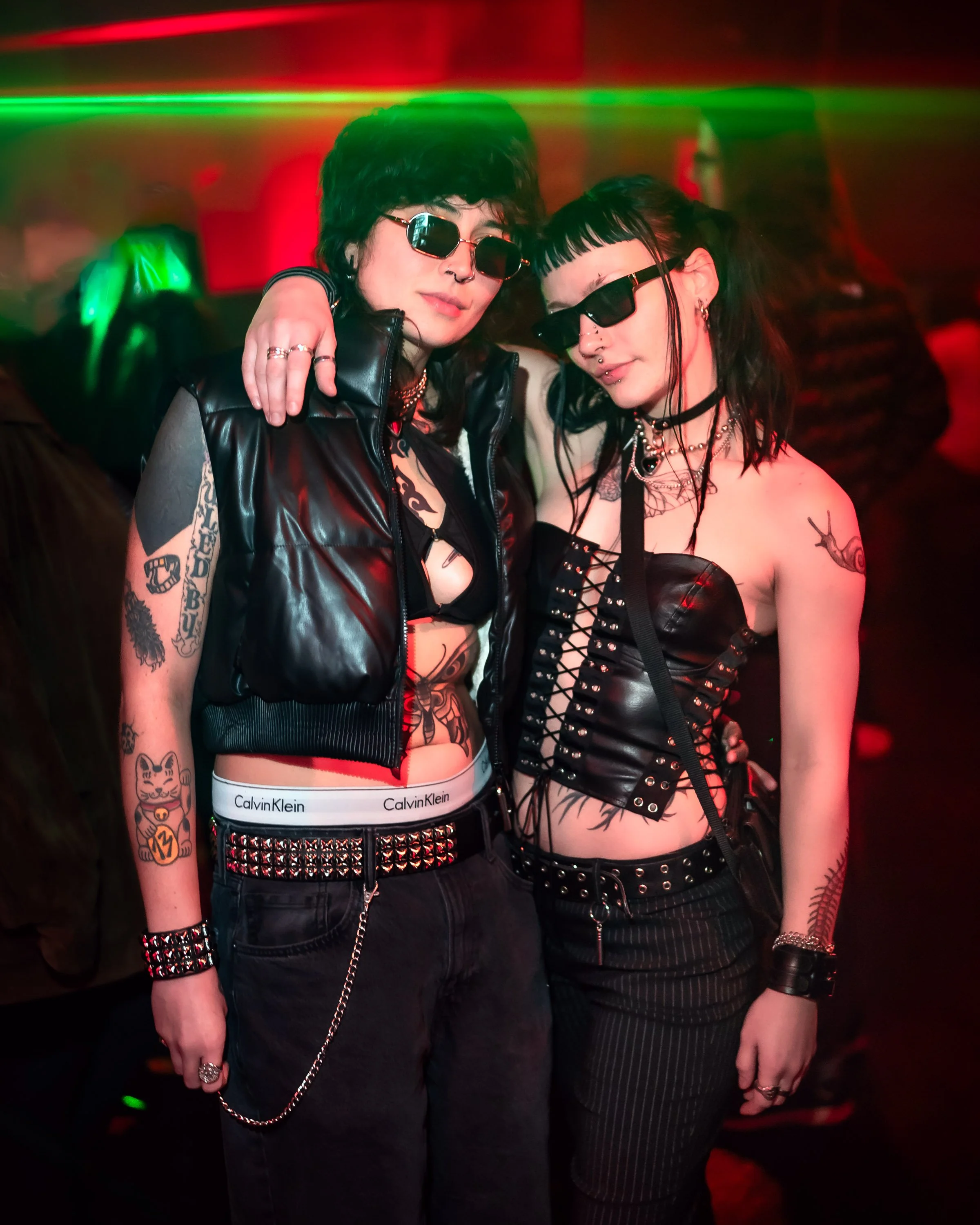 Two women in a nightclub wearing black gothic punk outfits, sunglasses, and displaying tattoos, standing closely together and posing for the camera.