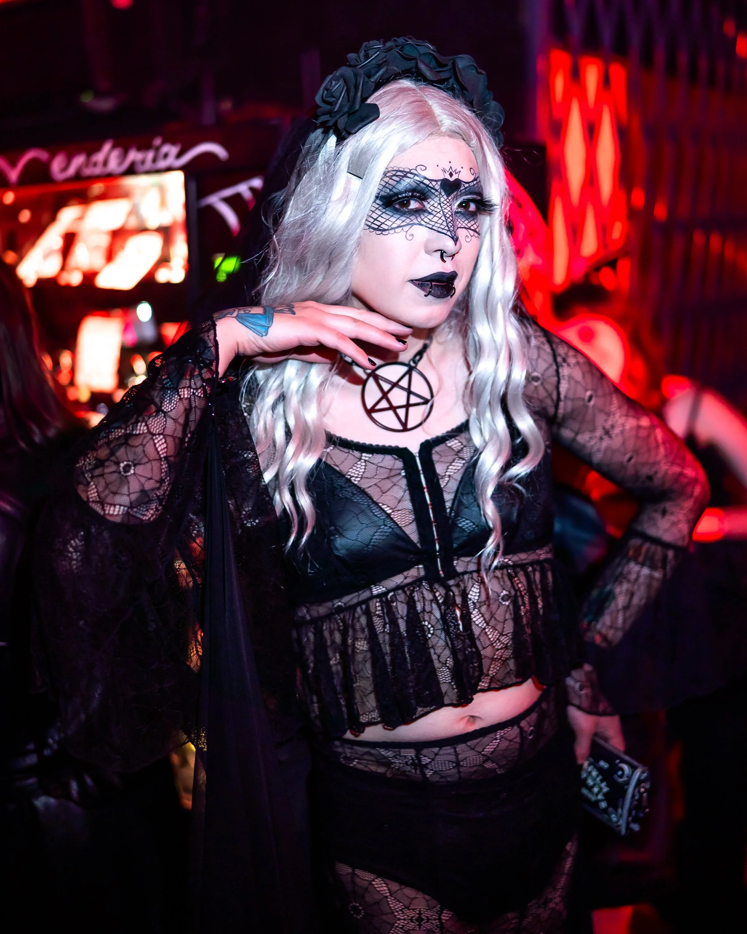 Woman dressed in gothic style with white wavy hair, black lace veil, and pentagram necklace, at a dark, red-lit party or club.