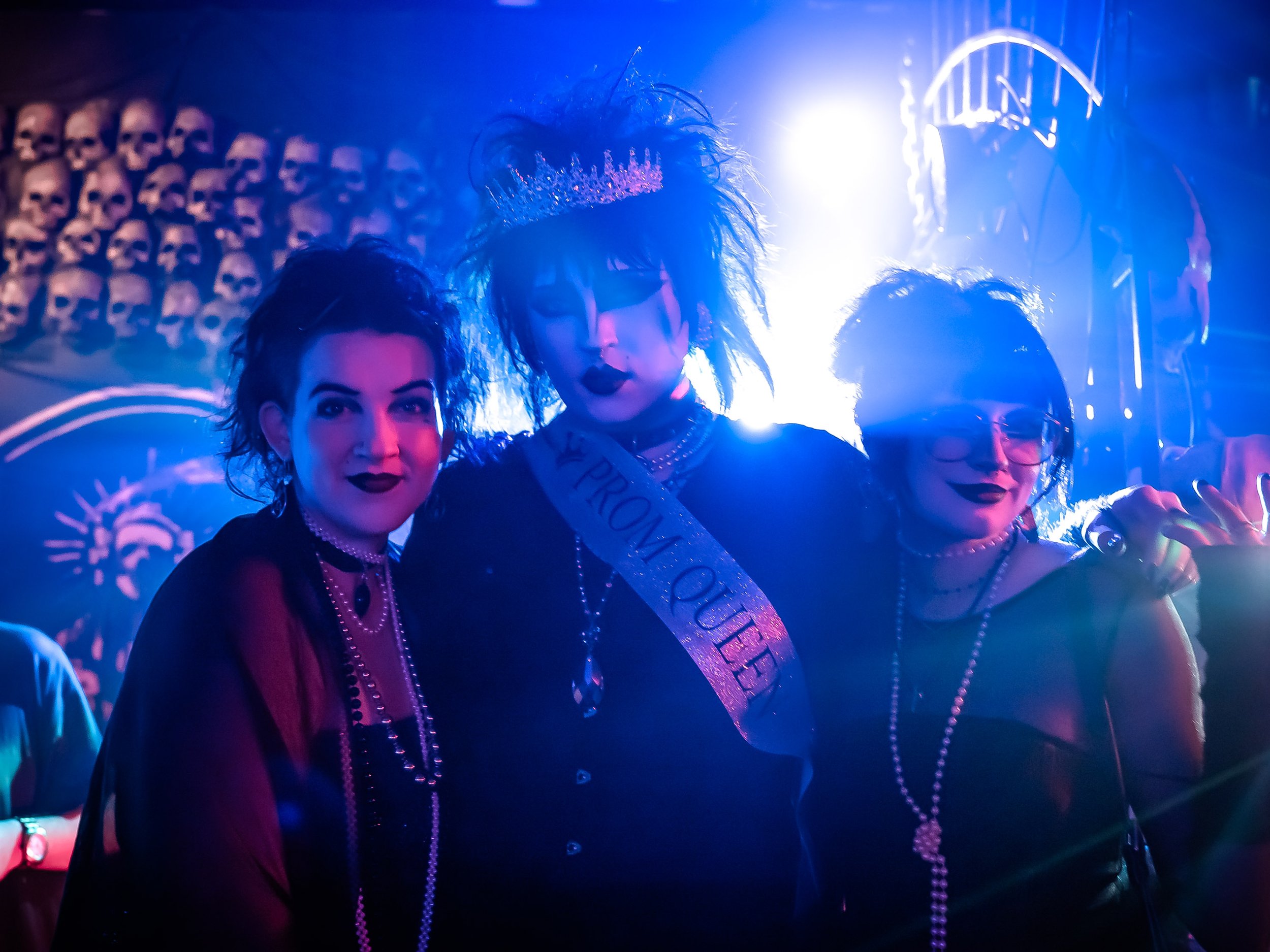 Three women dressed in punk/goth fashion with dark makeup and jewelry, posing together at a party or event with a colorful, neon-lit background.