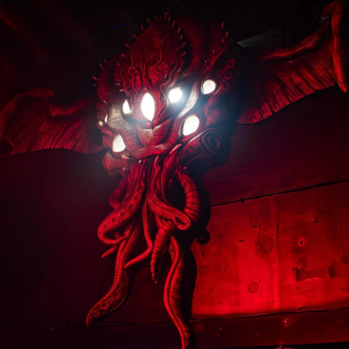 Large red and black creature with multiple glowing eyes and tentacle-like limbs, set against a dark background.