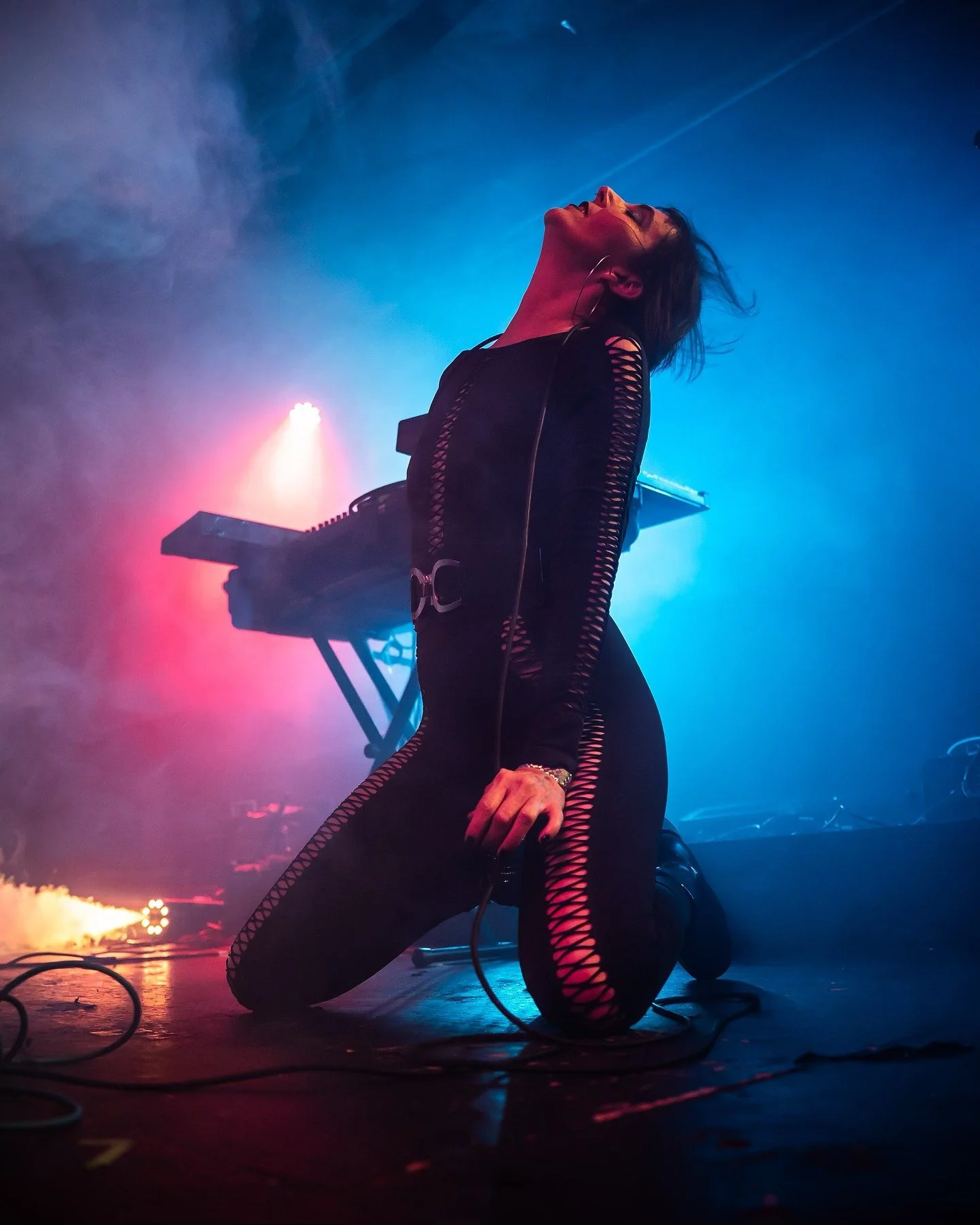 Performer kneeling on stage with head tilted back, surrounded by colorful stage lights, wearing a black outfit with cutouts, with musical equipment in the background.