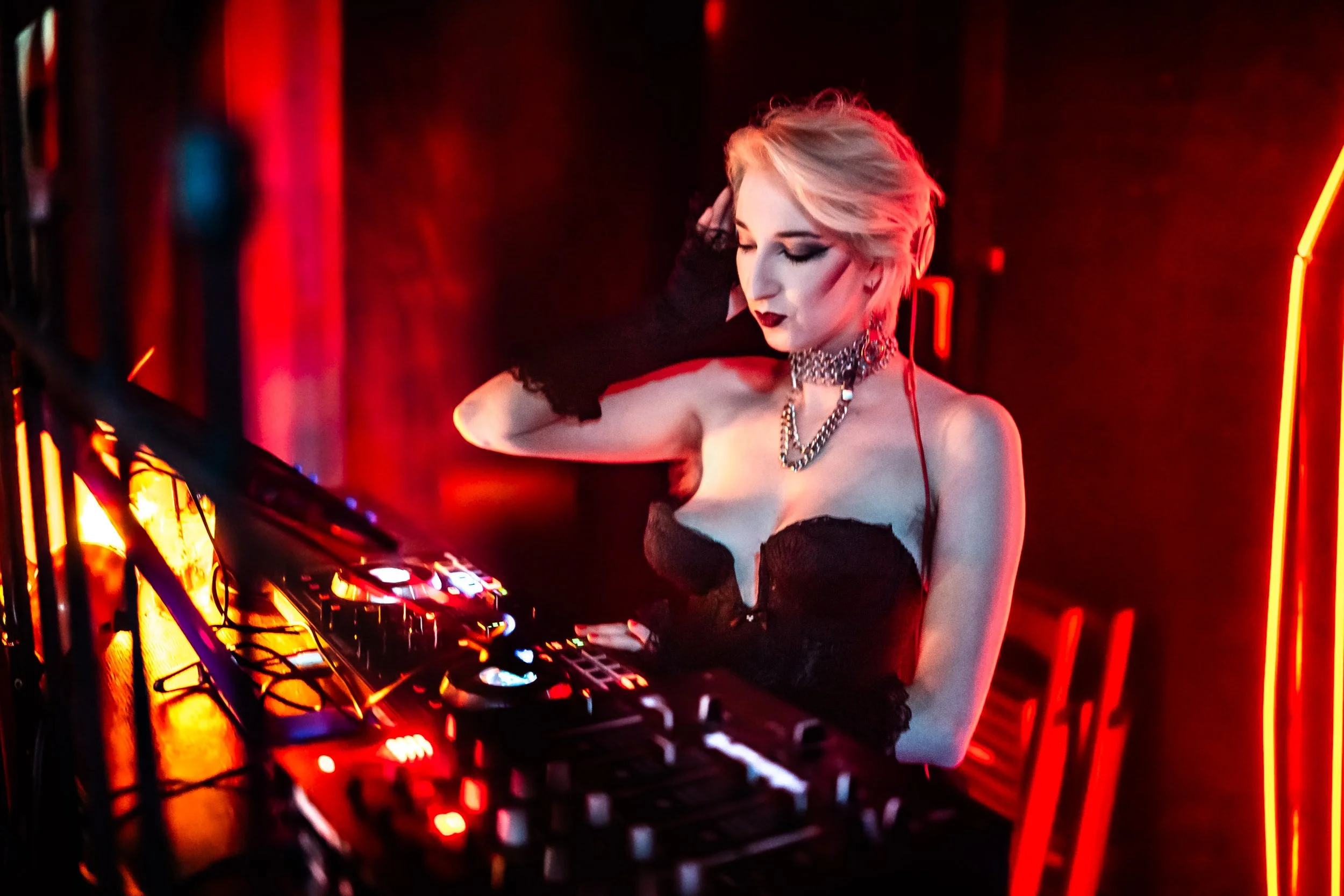 A woman with short platinum blonde hair, wearing a black lace corset and jewelry, stands behind DJ equipment in a dark, red-lit club or nightclub environment.