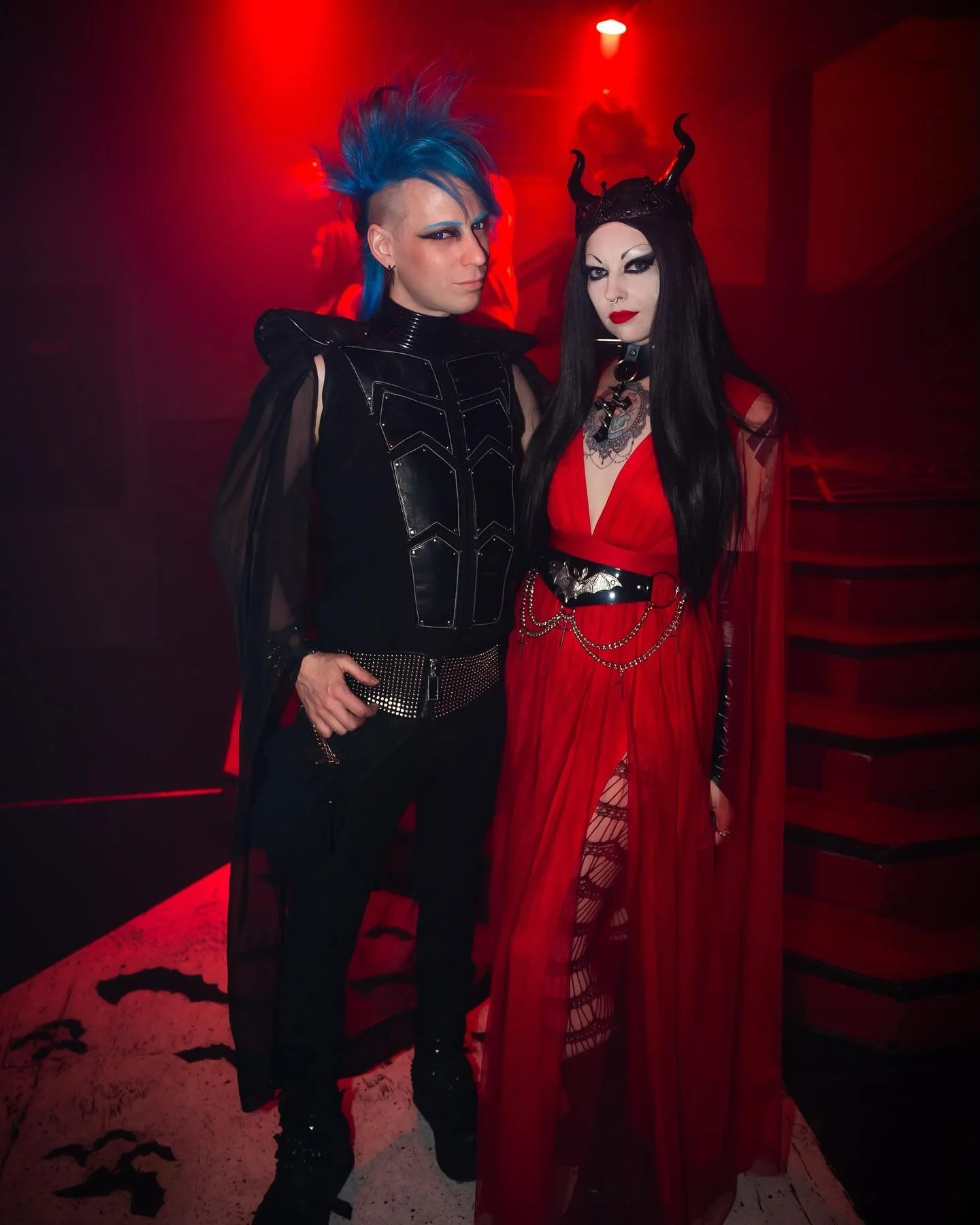 Two people in gothic costumes standing together in a dimly lit, red-tinged room. The person on the left has blue spiked hair, pale makeup, and is wearing a black leather and mesh outfit. The person on the right has long black hair, horns on their hea
