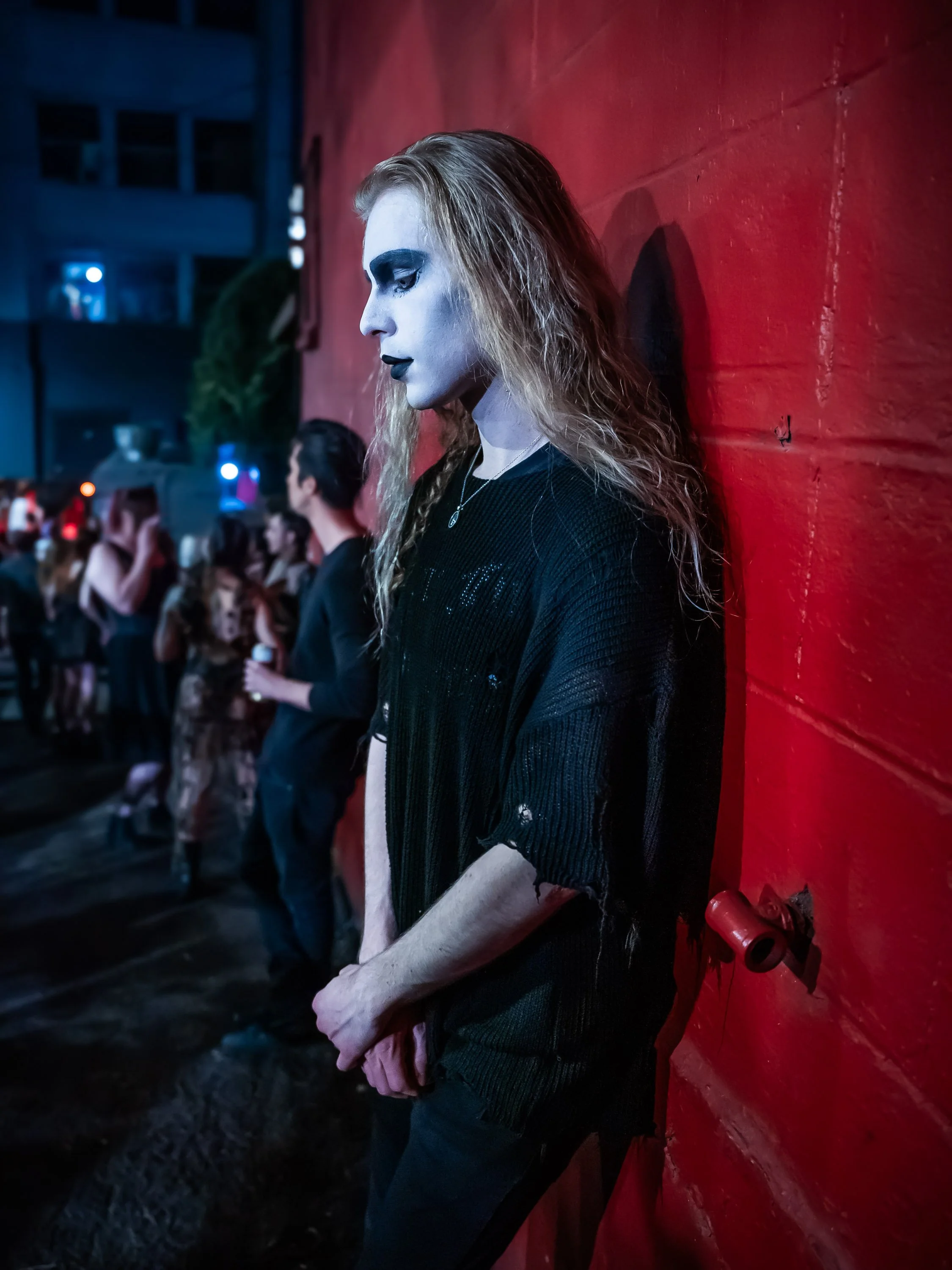 A woman with long curly hair and goth makeup, wearing black clothing, leaning against a red brick wall at a nighttime event with a crowd in the background.