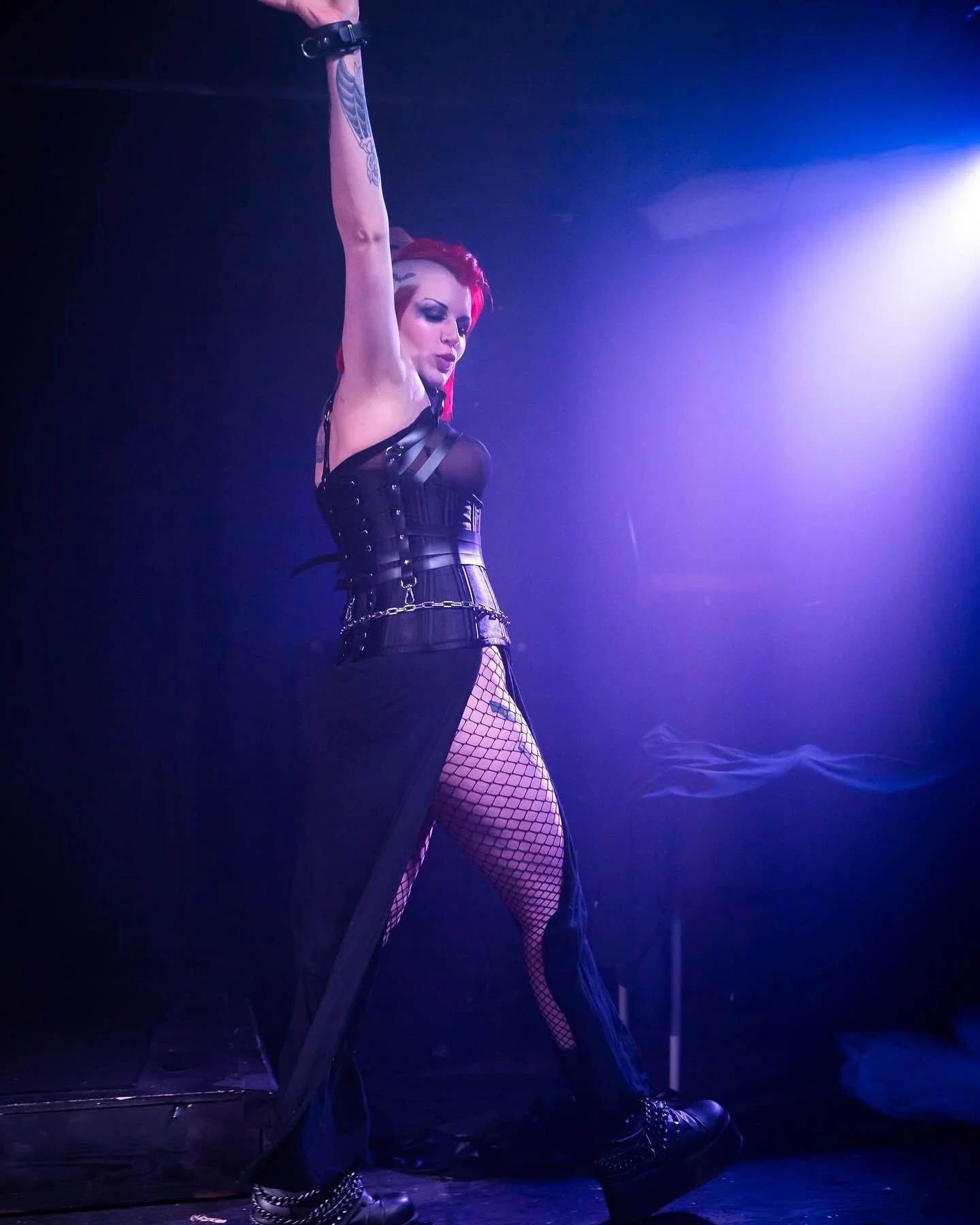 A woman with red hair and tattoos performing on stage with purple lighting, wearing black fishnet stockings, a black corset, and platform shoes.
