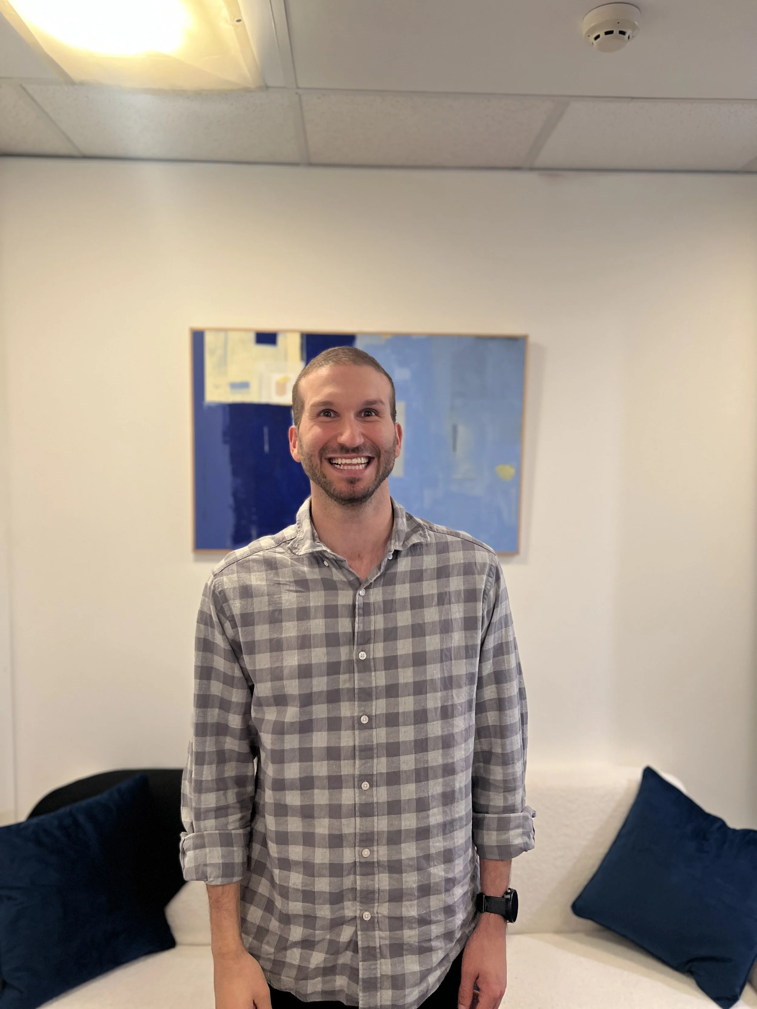A smiling man with short hair and a beard wearing a plaid shirt standing in a room with white walls and blue pillows, with a piece of abstract art on the wall behind him.