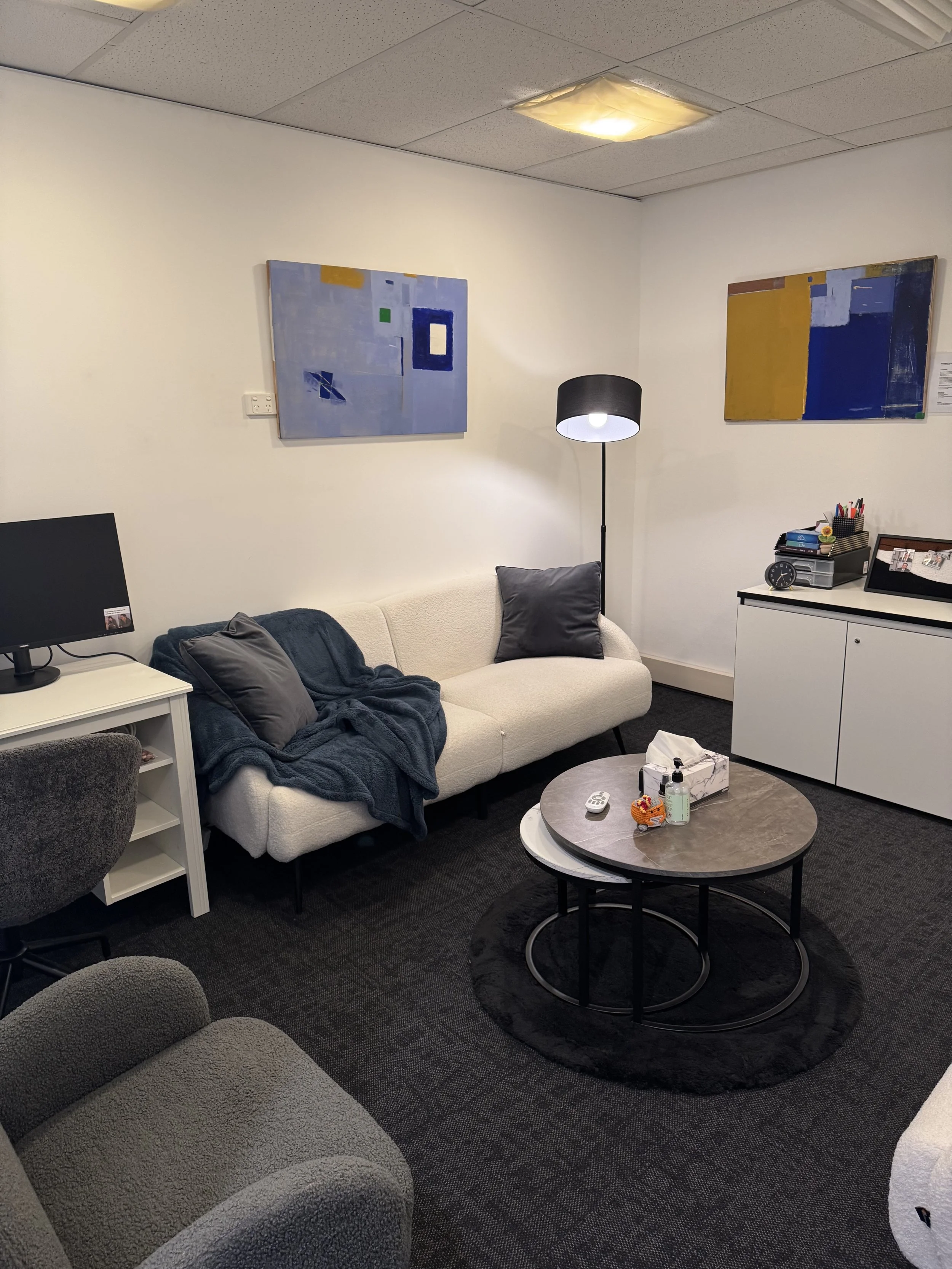 A cozy living room with a white couch adorned with gray and black pillows, a black floor lamp, a coffee table with a tissue box, hand sanitizer, and decorative items, a dark carpet, and wall art featuring abstract paintings in blue, gold, and white hues.