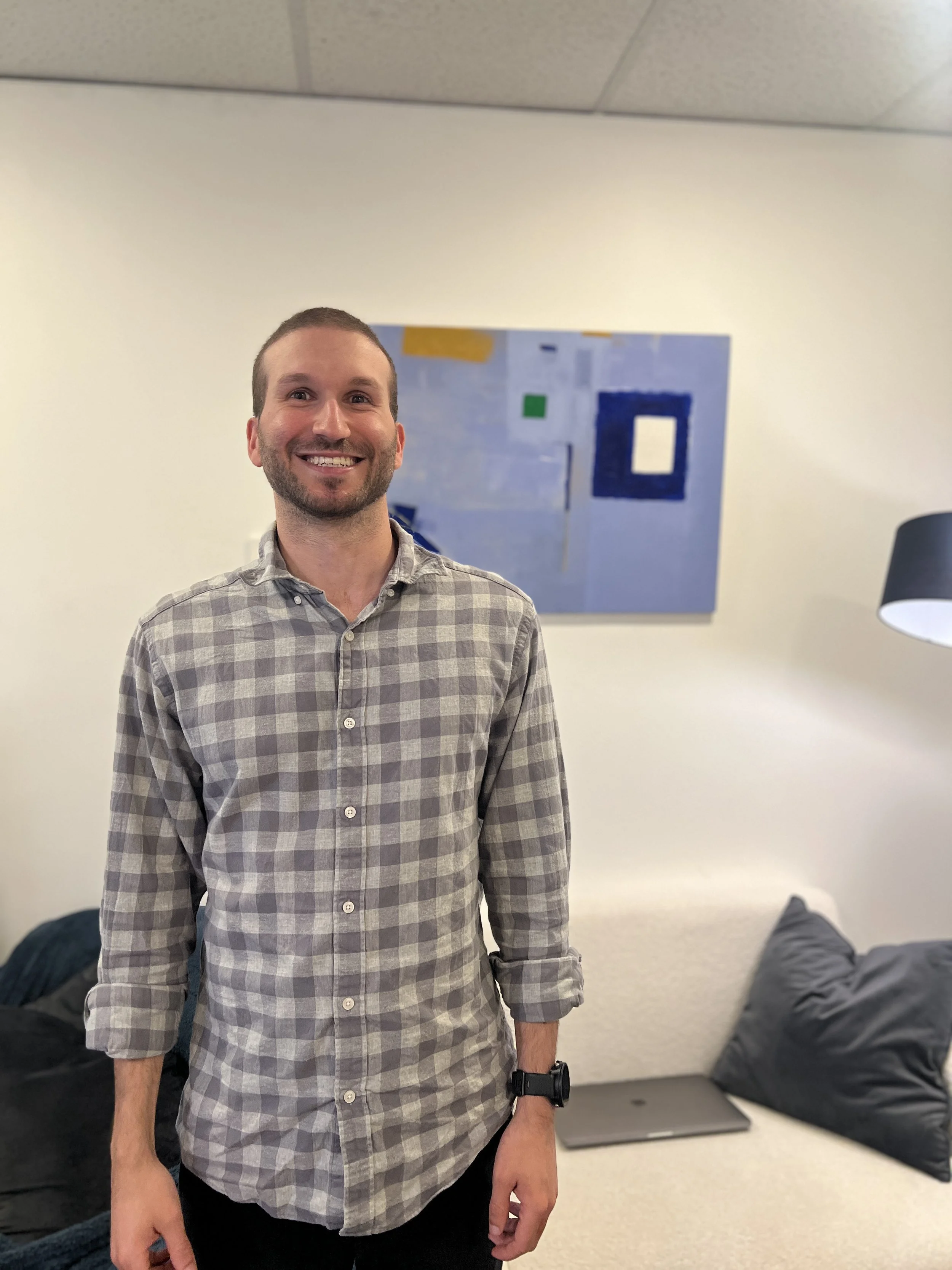 A man smiling, wearing a plaid shirt and a smartwatch, standing in an office with abstract art on the wall, a couch with pillows, and a laptop on a table.